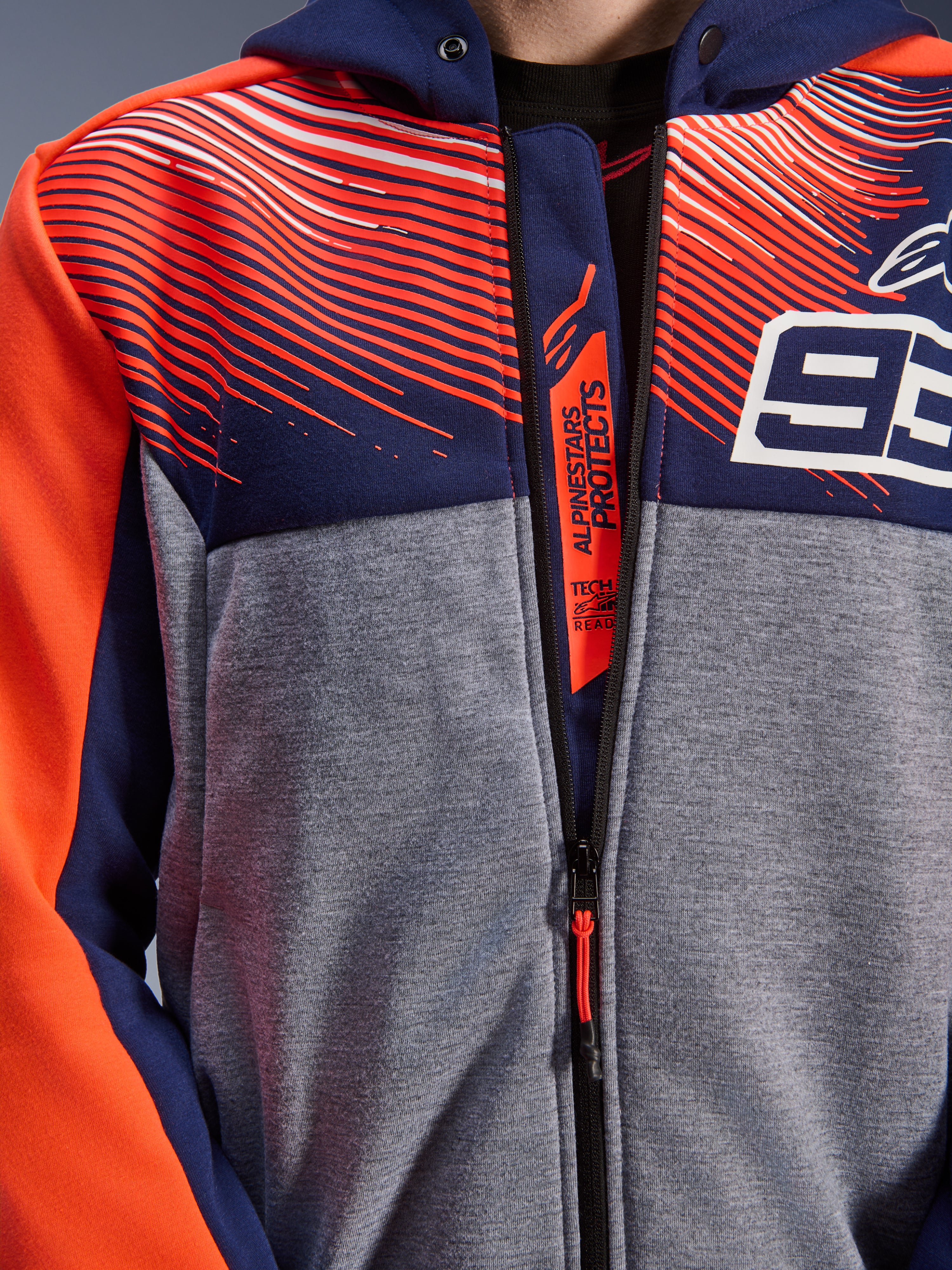 Alpinestars MM93 Chrome V2 Sport Hoodie, zbliżenie na klatkę piersiową i kołnierz, granatowy i szary melanżowy polar techniczny z czerwonymi grafikami fluo, białe logo 93, branding Alpinestars Protects na wewnętrznej patce zamka, odzież motocyklowa Tech-Air Ready