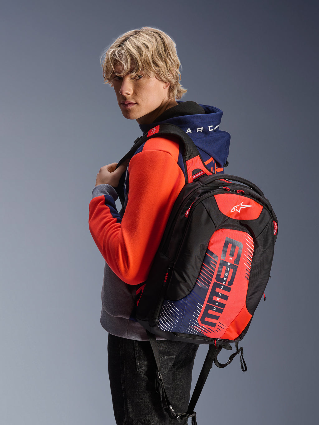 Męski model ubrany w bluzę z kapturem Alpinestars MM93 Chrome V2 Sport w kolorach Blue Navy Gray Mel i Red Fluo, w zestawie z dopasowanym plecakiem z logo MM93, miejska odzież motocyklowa o nowoczesnej sportowej sylwetce.