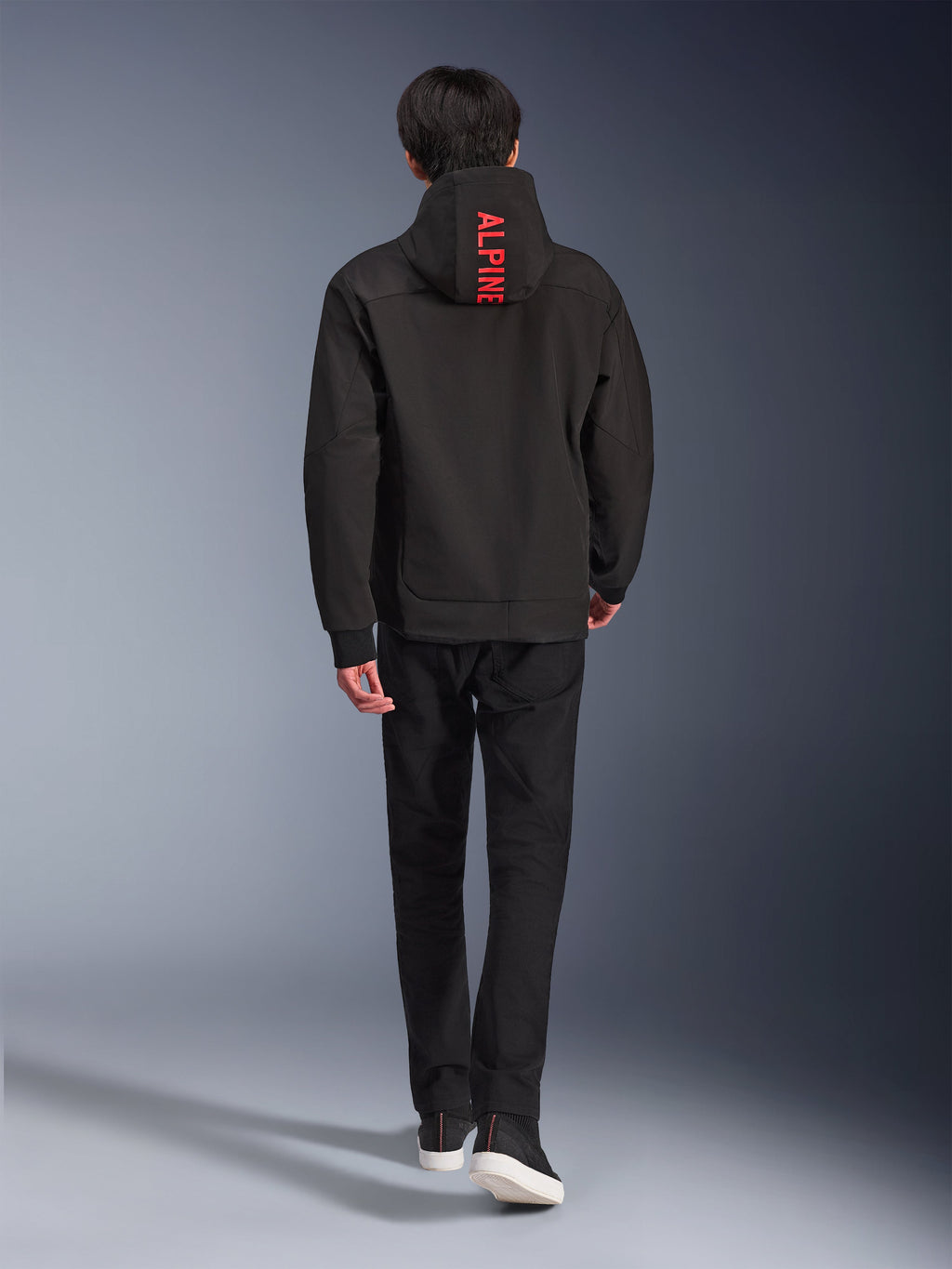 Alpinestars Radium Tech Hoodie, miejska kurtka motocyklowa, czarna, widok z tyłu z jasnoczerwonym napisem „ALPINE” na kapturze, noszona z czarnymi spodniami i sneakersami, stylowa techniczna konstrukcja typu shell do codziennych dojazdów.