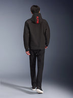 Alpinestars Radium Tech Hoodie, miejska kurtka motocyklowa, czarna, widok z tyłu z jasnoczerwonym napisem „ALPINE” na kapturze, noszona z czarnymi spodniami i sneakersami, stylowa techniczna konstrukcja typu shell do codziennych dojazdów.