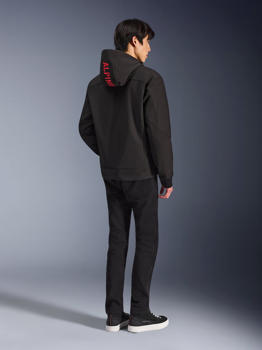 Alpinestars Radium Tech Hoodie, czarna bluza z czerwonym brandingiem Alpinestars na kapturze, widok z tyłu na modelu, noszona z czarnymi spodniami i tenisówkami, techniczna miejska odzież motocyklowa