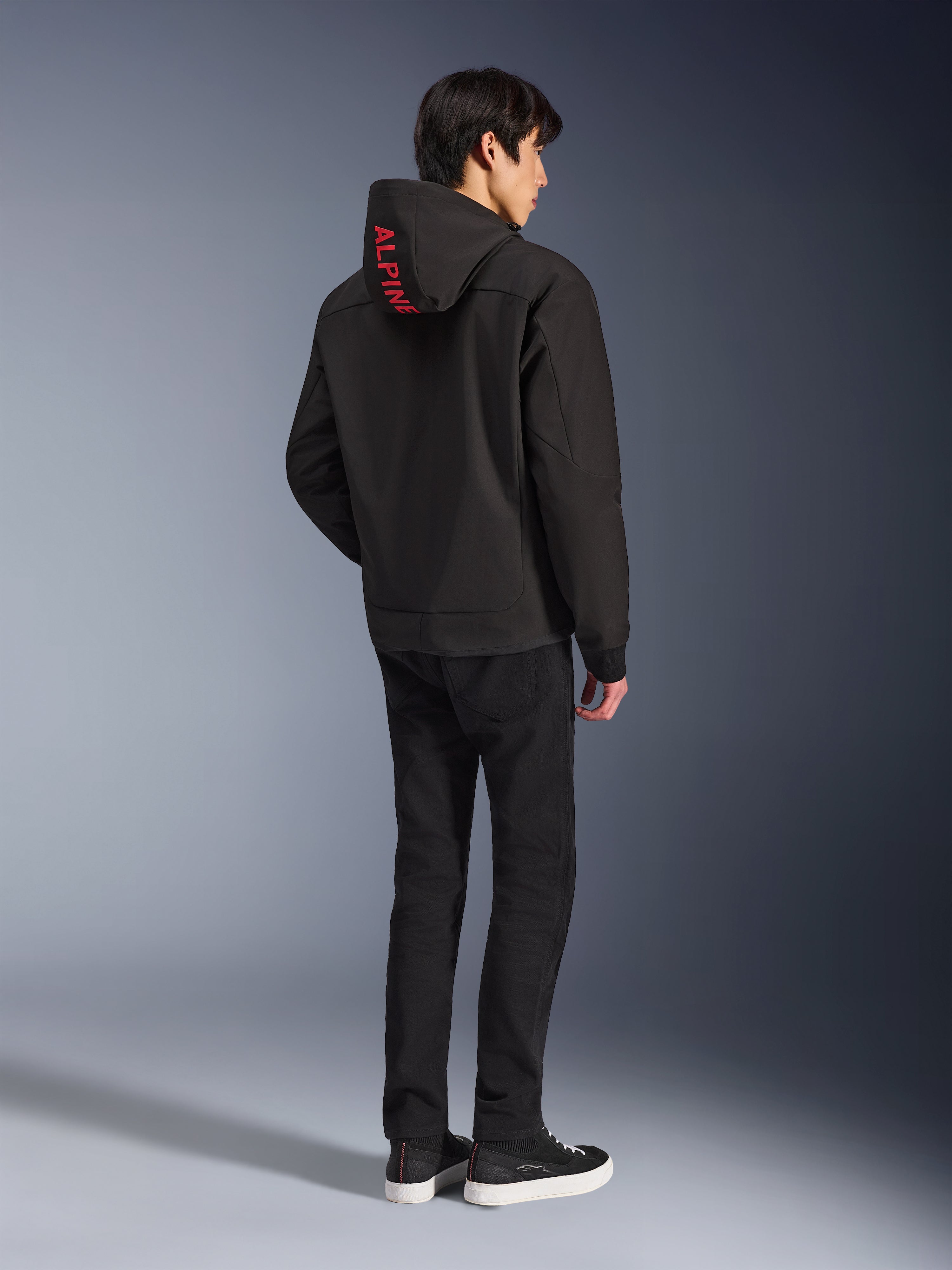 Alpinestars Radium Tech Hoodie, czarna bluza z czerwonym brandingiem Alpinestars na kapturze, widok z tyłu na modelu, noszona z czarnymi spodniami i tenisówkami, techniczna miejska odzież motocyklowa