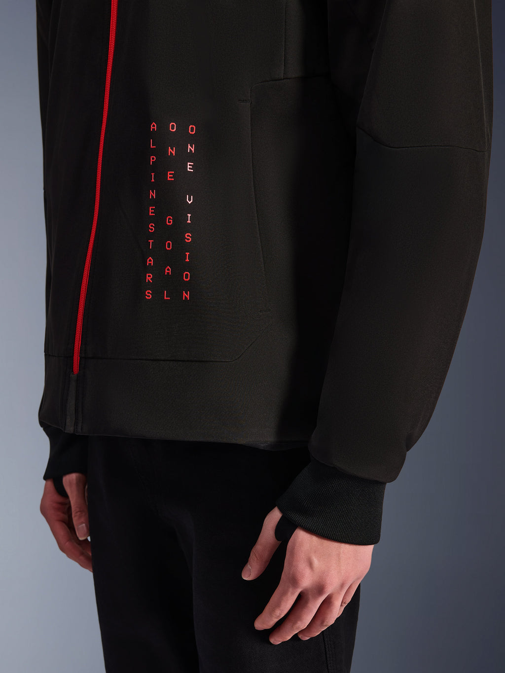 Alpinestars Radium Tech Hoodie, miejska kurtka motocyklowa, czarna z jaskrawoczerwonymi detalami, zbliżenie na grafikę z przodu z pionowym czerwonym napisem „ALPINESTARS ONE GOAL ONE VISION”, czerwony zamek błyskawiczny i prążkowany mankiet z otworem na kciuk