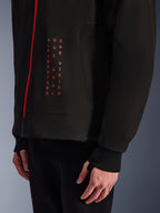 Alpinestars Radium Tech Hoodie, miejska kurtka motocyklowa, czarna z jaskrawoczerwonymi detalami, zbliżenie na grafikę z przodu z pionowym czerwonym napisem „ALPINESTARS ONE GOAL ONE VISION”, czerwony zamek błyskawiczny i prążkowany mankiet z otworem na kciuk