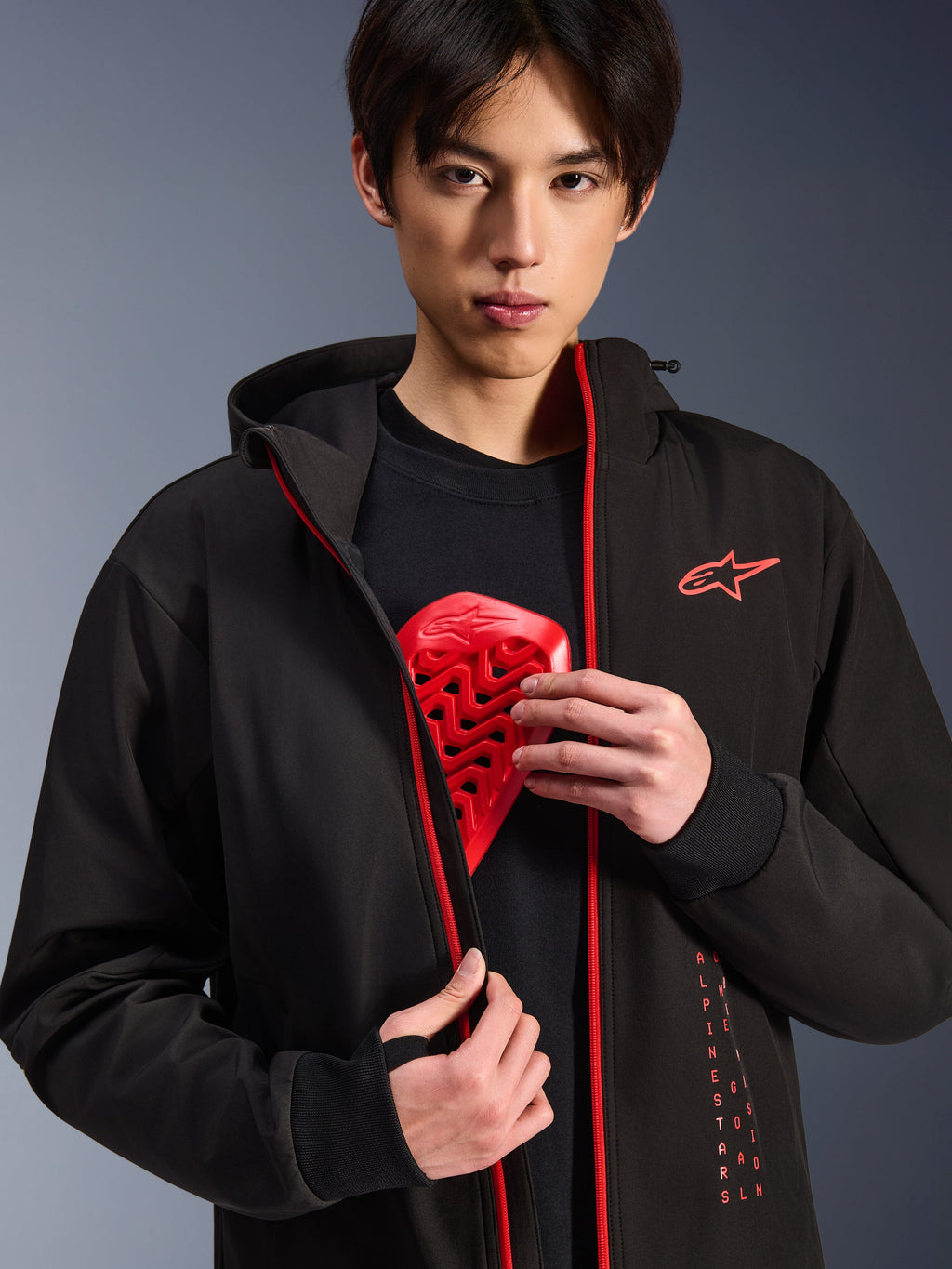 Alpinestars Radium Tech Hoodie, miejska kurtka motocyklowa, czarna z jaskrawoczerwonymi elementami, model prezentujący czerwoną wkładkę ochraniacza klatki piersiowej, jaskrawoczerwone zamki i pionowe logo Alpinestars, wiatroszczelna powłoka techniczna do jazdy miejskiej