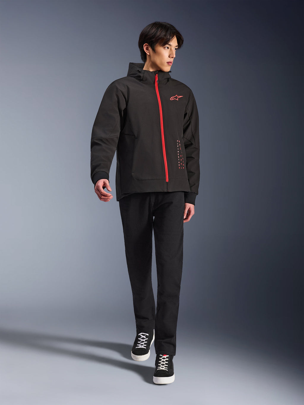 Alpinestars Radium Tech Hoodie w kolorze Black Bright Red, miejska kurtka motocyklowa noszona przez modela, z czerwonym przednim zamkiem i logo na piersi, stylizowana z czarnymi spodniami i sneakersami dla nowoczesnego, miejskiego looku.