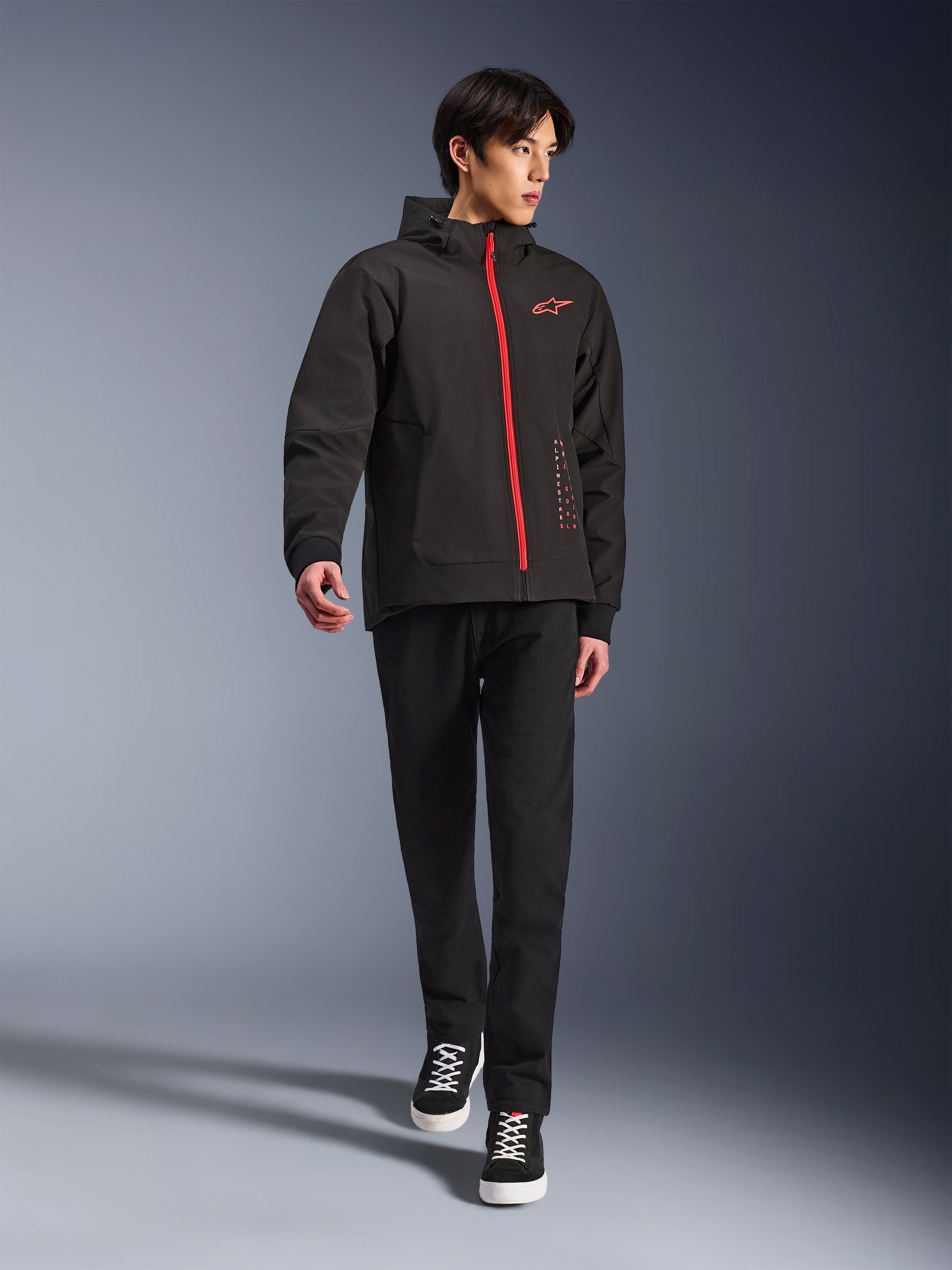Alpinestars Radium Tech Hoodie w kolorze Black Bright Red, miejska kurtka motocyklowa noszona przez modela, z czerwonym przednim zamkiem i logo na piersi, stylizowana z czarnymi spodniami i sneakersami dla nowoczesnego, miejskiego looku.