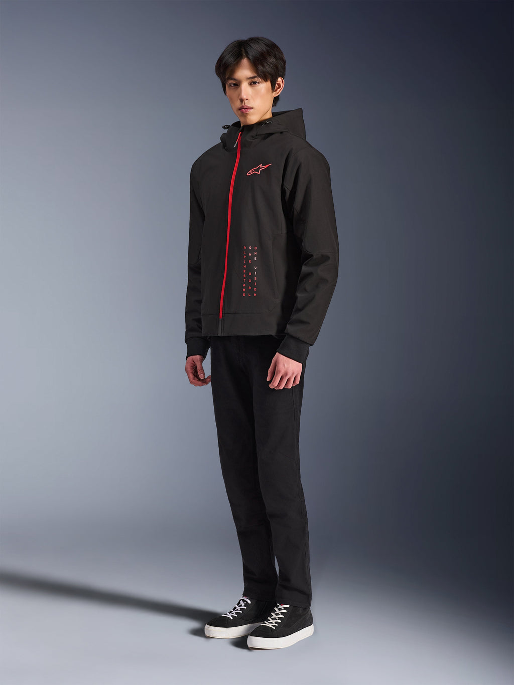 Alpinestars Radium Tech Hoodie, techniczna bluza motocyklowa do jazdy miejskiej, czarna z jasnoczerwonymi elementami, czerwony zamek błyskawiczny i logo na piersi, model w czarnych spodniach i czarnych trampkach z białą podeszwą, studyjne ujęcie całej sylwetki
