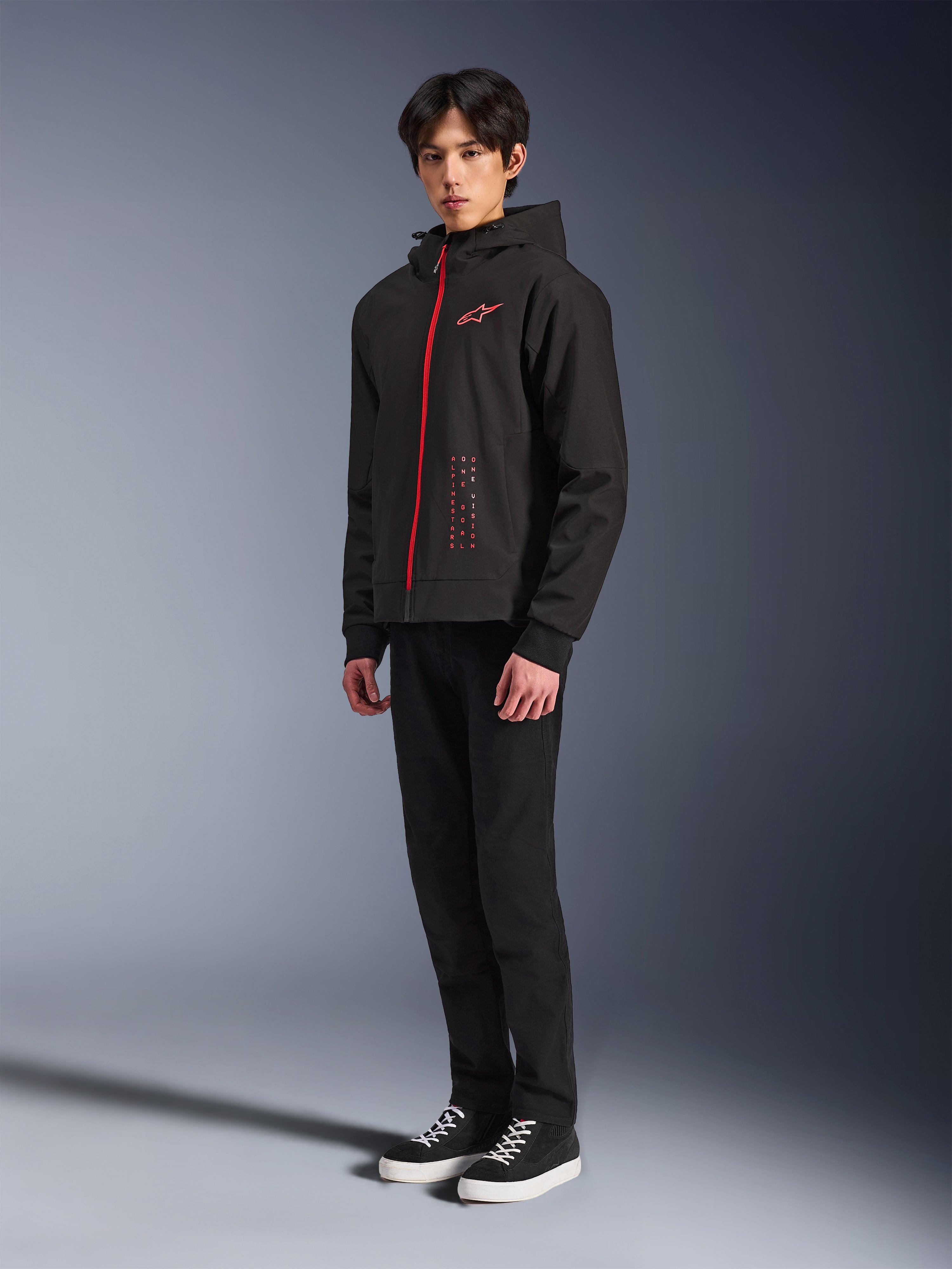 Alpinestars Radium Tech Hoodie, techniczna bluza motocyklowa do jazdy miejskiej, czarna z jasnoczerwonymi elementami, czerwony zamek błyskawiczny i logo na piersi, model w czarnych spodniach i czarnych trampkach z białą podeszwą, studyjne ujęcie całej sylwetki