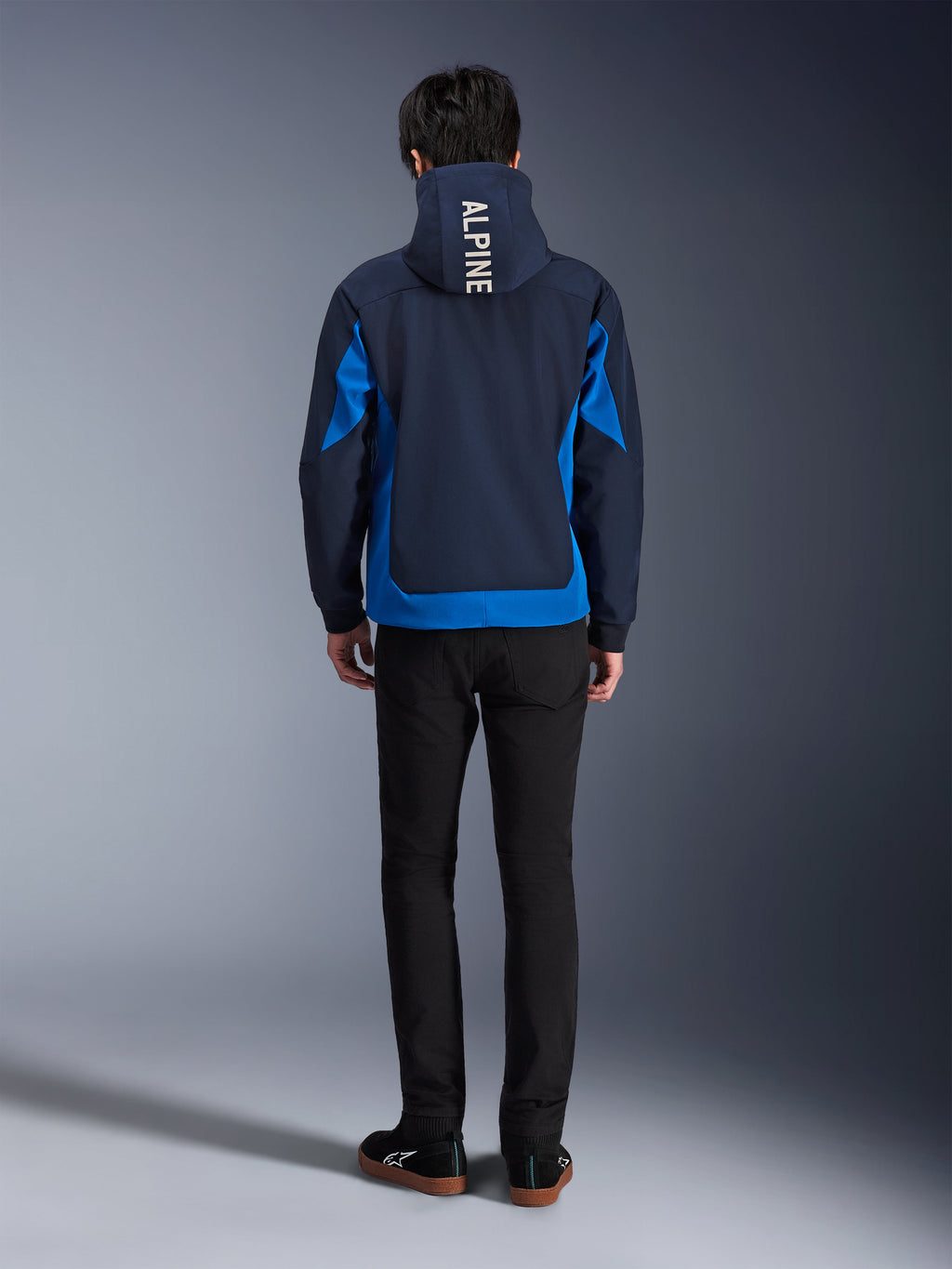 Alpinestars Radium Tech Hoodie, kolor Night Navy i Royal Blue, widok z tyłu z pionowym napisem „ALPINE” na kapturze, niebieskie panele boczne i dolne wykończenie, casualowa miejska bluza motocyklowa z wiatroszczelną powłoką 3L, noszona z czarnymi spodniami