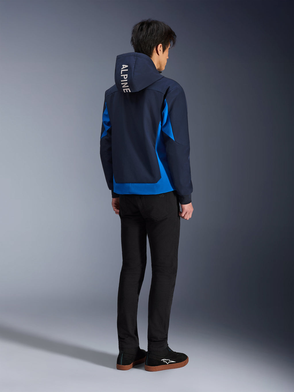 Alpinestars Radium Tech Hoodie, Night Navy i Royal Blue, widok z tyłu modela w technicznej bluzie z kapturem z białym napisem „ALPINE” na kapturze, w zestawie z czarnymi spodniami i butami, miejska odzież motocyklowa