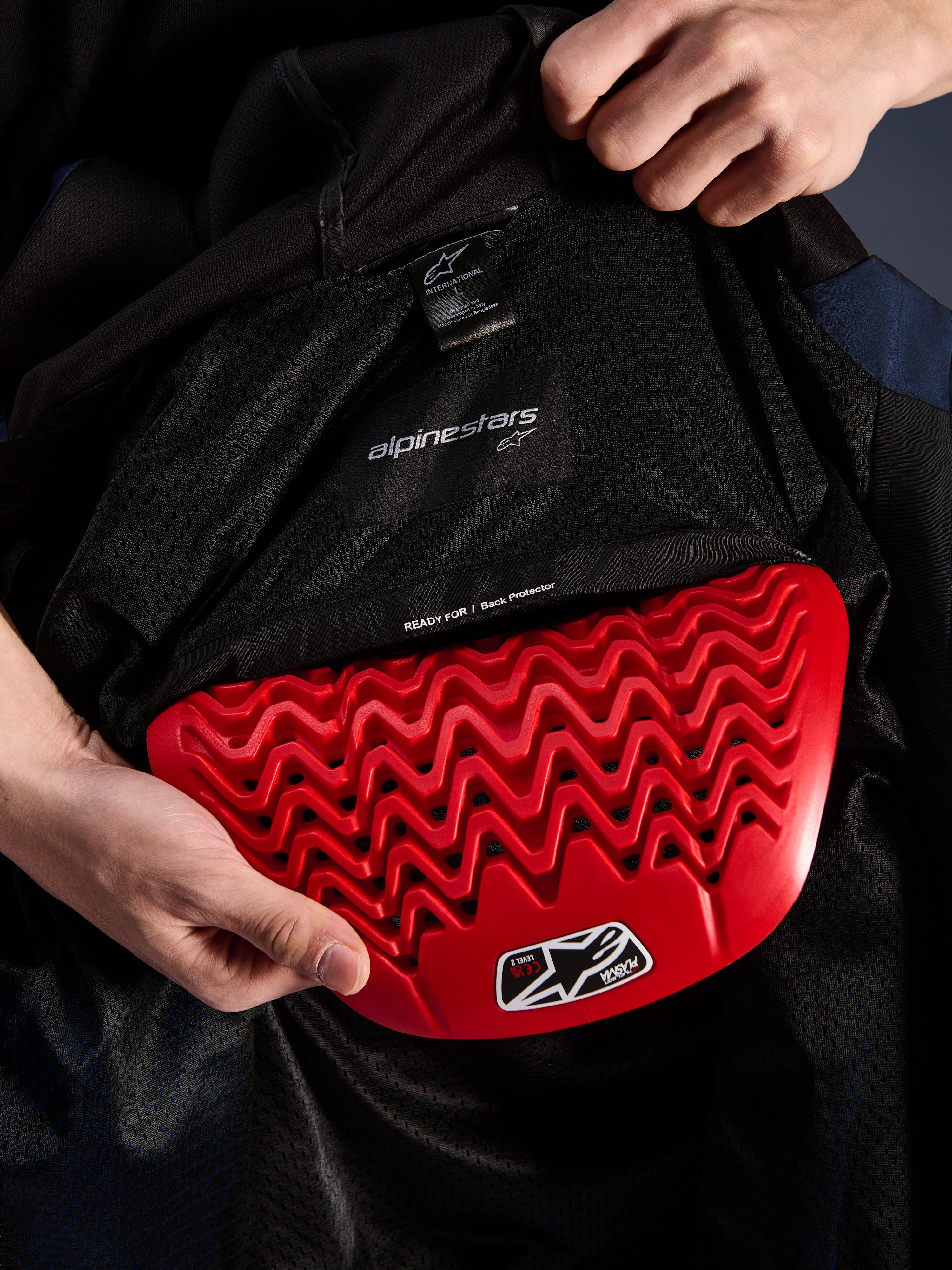 Alpinestars Radium Tech Hoodie, detal wnętrza, czerwony ochraniacz pleców Nucleon Plasma wkładany do kieszeni na ochraniacz, czarna podszewka mesh z napisem 'READY FOR | Back Protector', falista struktura plastra miodu, techniczne elementy ochronne miejskiej kurtki motocyklowej