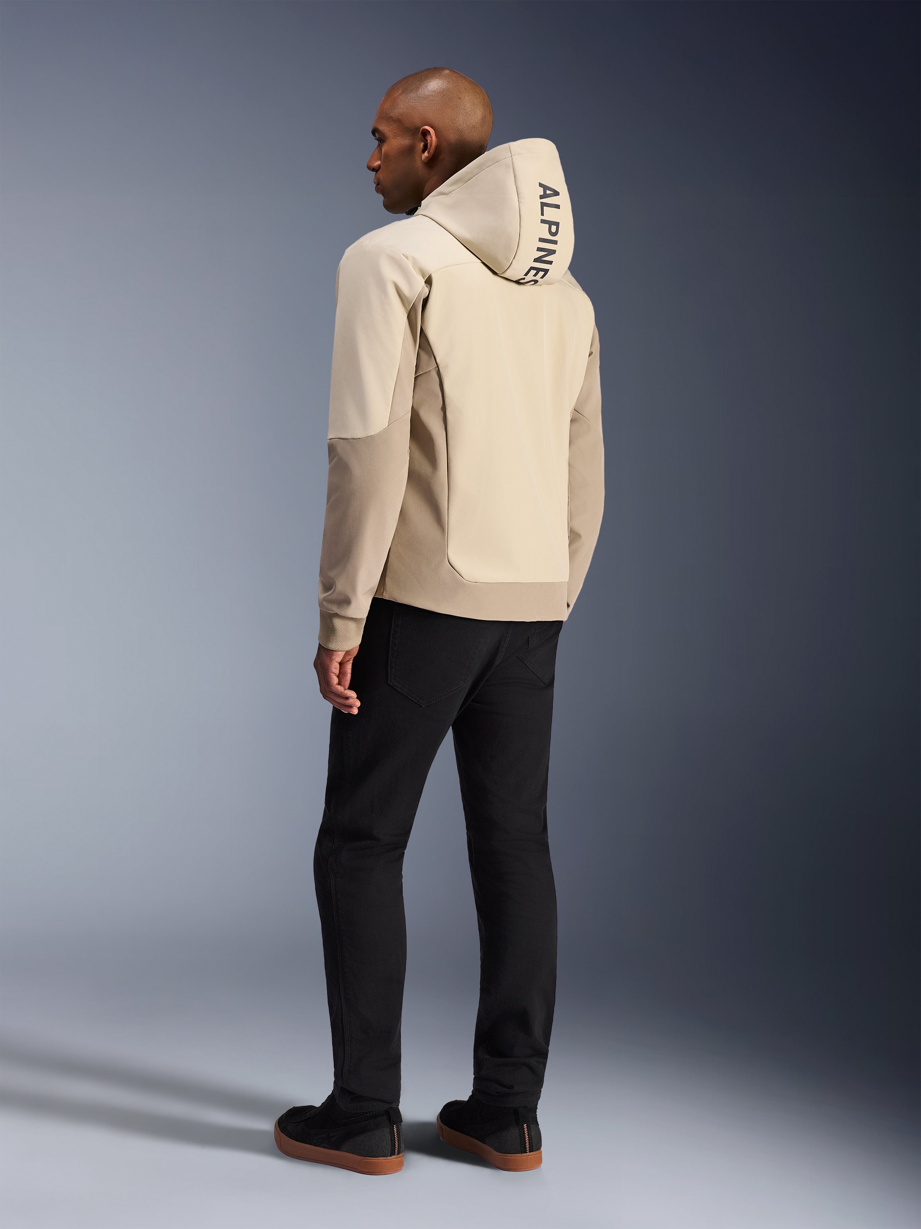 Alpinestars Radium Tech Hoodie w kolorze Mastic Aluminum, miejska kurtka motocyklowa, widok z tyłu na modelu z założonym kapturem, pionowy napis ALPINESTARS na kapturze, w zestawieniu z czarnymi spodniami, nowoczesna tekstylna konstrukcja zapewniająca ochronę podczas jazdy miejskiej