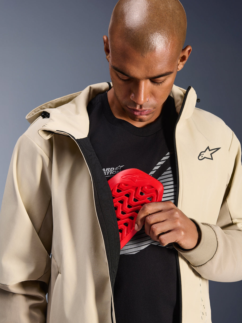 Alpinestars Radium Tech Hoodie w kolorze Mastic Aluminum, miejska kurtka motocyklowa, model prezentujący wewnętrzną kieszeń na ochraniacz z czerwonym ochraniaczem klatki piersiowej Nucleon, wiatroszczelna powłoka techniczna o fasonie codziennej bluzy z kapturem z brandingiem Alpinestars