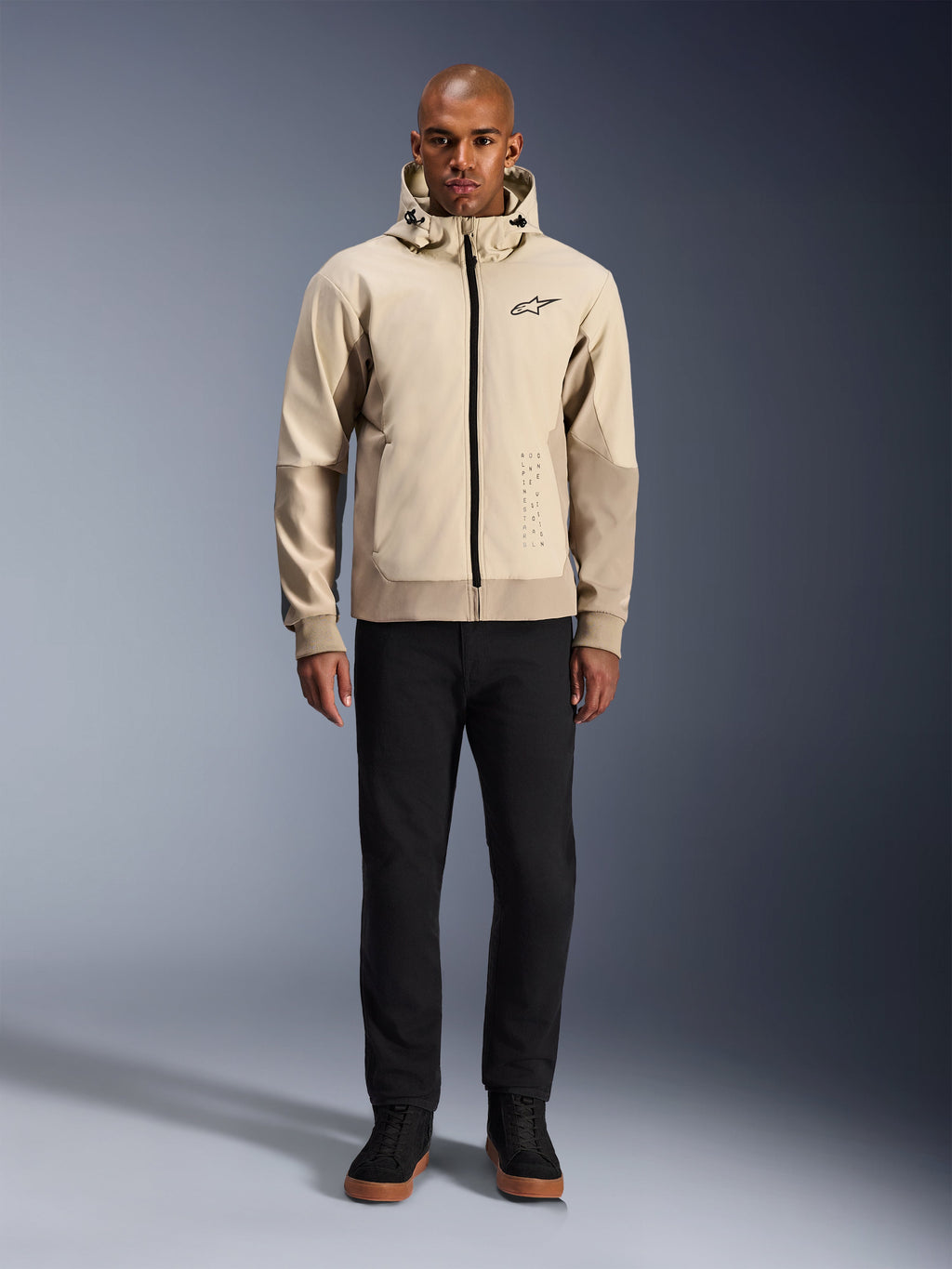 Alpinestars Radium Tech Hoodie w kolorze Mastic Aluminum, miejska bluza motocyklowa prezentowana na modelu w czarnych spodniach i butach, z czarnym zamkiem błyskawicznym na całej długości, małym logo na piersi, o sportowym kroju do jazdy miejskiej