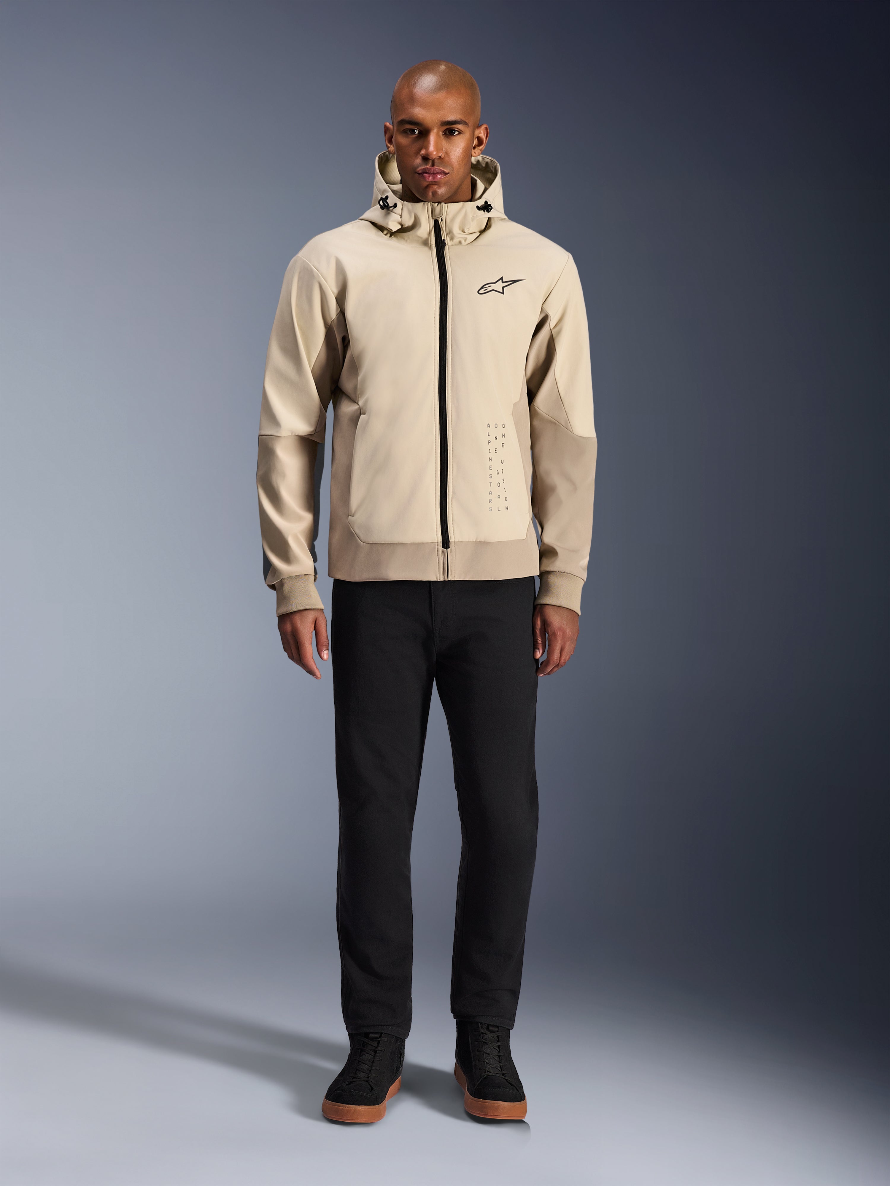Alpinestars Radium Tech Hoodie w kolorze Mastic Aluminum, miejska bluza motocyklowa prezentowana na modelu w czarnych spodniach i butach, z czarnym zamkiem błyskawicznym na całej długości, małym logo na piersi, o sportowym kroju do jazdy miejskiej