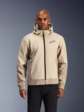 Alpinestars Radium Tech Hoodie w kolorze Mastic Aluminum, jasnobeżowa i piaskowa miejska kurtka motocyklowa, czarny zamek błyskawiczny z przodu i małe logo na klatce piersiowej, prezentowana przez mężczyznę w czarnych spodniach, techniczna wiatroszczelna powłoka w nowoczesnej stylistyce streetwear