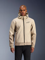 Alpinestars Radium Tech Hoodie w kolorze Mastic Aluminum, jasnobeżowa i piaskowa miejska kurtka motocyklowa, czarny zamek błyskawiczny z przodu i małe logo na klatce piersiowej, prezentowana przez mężczyznę w czarnych spodniach, techniczna wiatroszczelna powłoka w nowoczesnej stylistyce streetwear