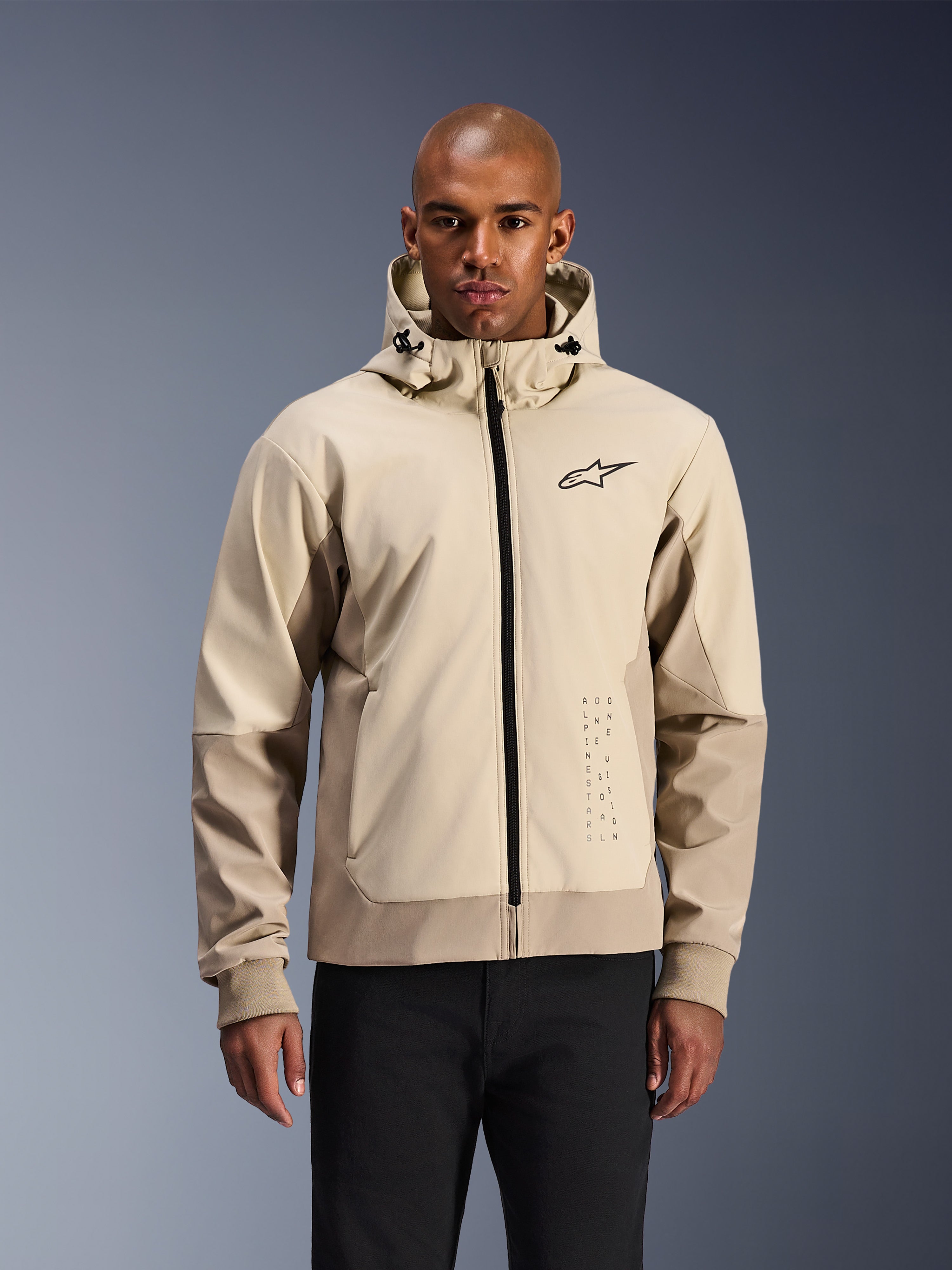 Alpinestars Radium Tech Hoodie w kolorze Mastic Aluminum, jasnobeżowa i piaskowa miejska kurtka motocyklowa, czarny zamek błyskawiczny z przodu i małe logo na klatce piersiowej, prezentowana przez mężczyznę w czarnych spodniach, techniczna wiatroszczelna powłoka w nowoczesnej stylistyce streetwear