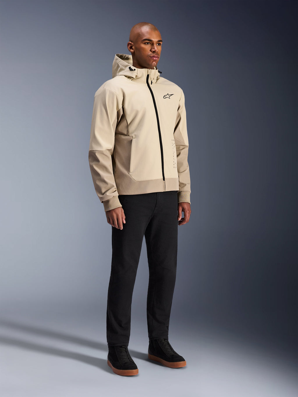 Alpinestars Radium Tech Hoodie, miejska kurtka motocyklowa, jasnobeżowy Mastic Aluminum, noszona z czarnymi spodniami i czarnymi wysokimi sneakersami, wyposażona w techniczną wiatroszczelną powłokę, przedni zamek błyskawiczny i odpinany kaptur, nowoczesny design streetwearowy do jazdy miejskiej