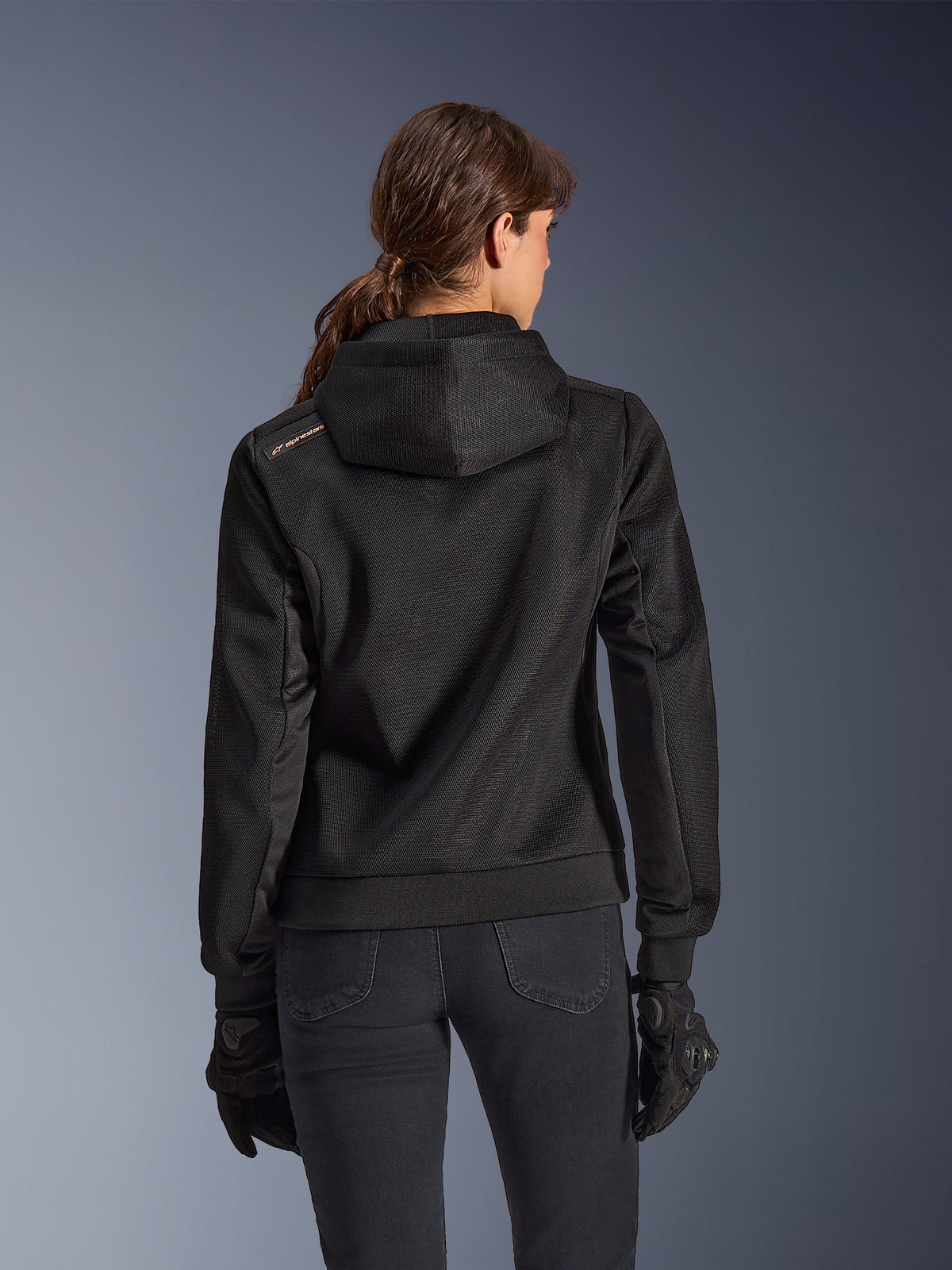 Alpinestars Stella Chrome Superairflow Hoodie, widok z tyłu kobiety w czarnej technicznej bluzie motocyklowej z siatki 3D, zaprojektowany kaptur i miedziane logo na ramieniu, czarne rękawice i spodnie motocyklowe