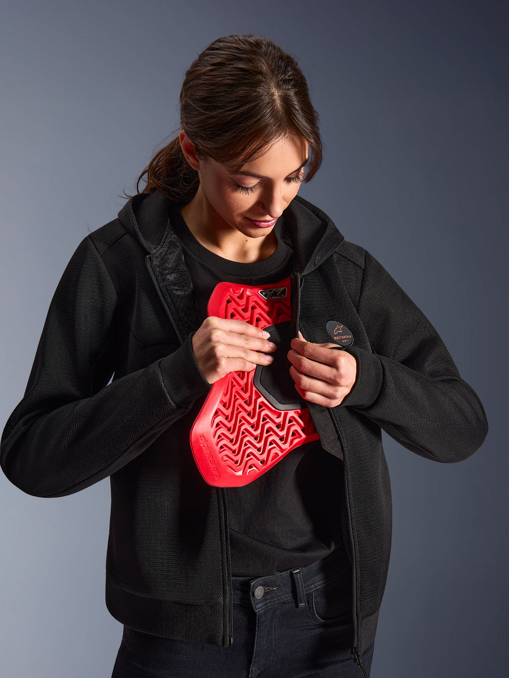 Alpinestars Stella Chrome Superairflow Hoodie, damska bluza motocyklowa, czarna, modelka trzymająca czerwony ochraniacz klatki piersiowej Nucleon, lekka konstrukcja z siatki 3D do jazdy miejskiej i sportowej