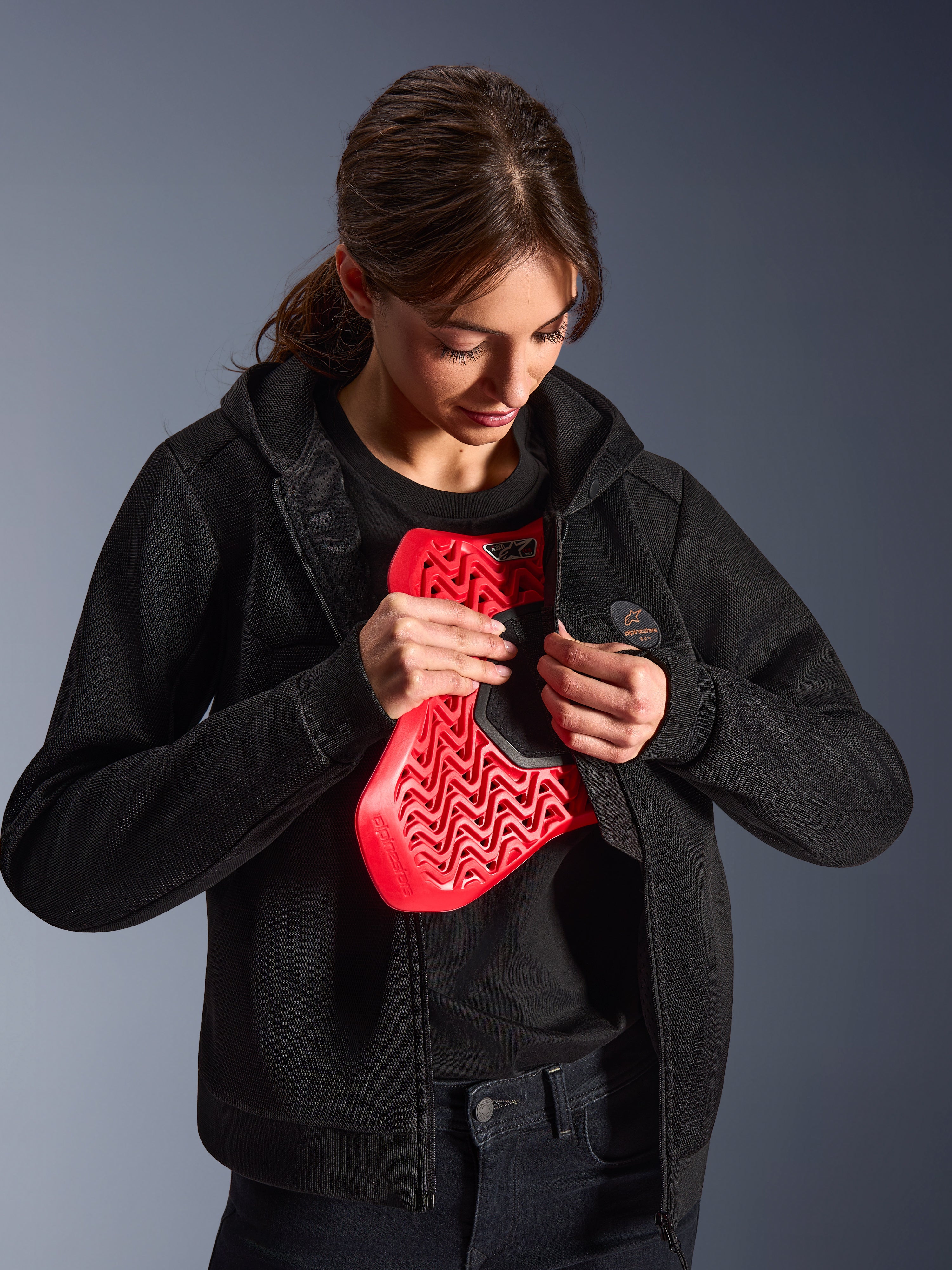 Alpinestars Stella Chrome Superairflow Hoodie, damska bluza motocyklowa, czarna, modelka trzymająca czerwony ochraniacz klatki piersiowej Nucleon, lekka konstrukcja z siatki 3D do jazdy miejskiej i sportowej