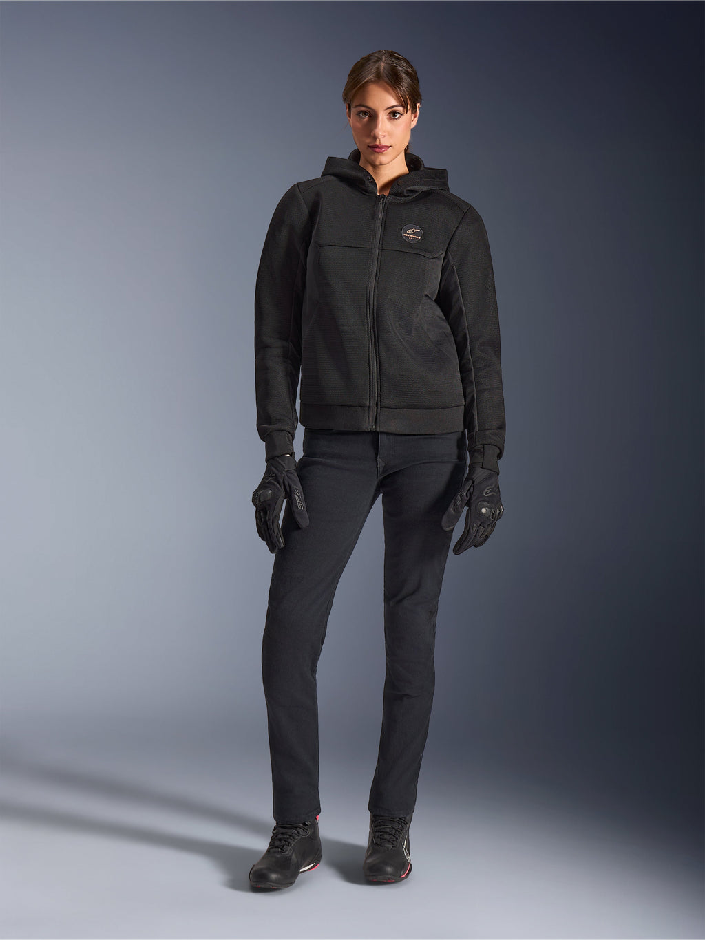 Modelka w bluzie z kapturem Alpinestars Stella Chrome Superairflow w kolorze czarnym, czarne techniczne spodnie do jazdy i rękawice, konstrukcja z siatki 3D, miedziane logo na piersi, miejska sportowa odzież motocyklowa dla kobiet