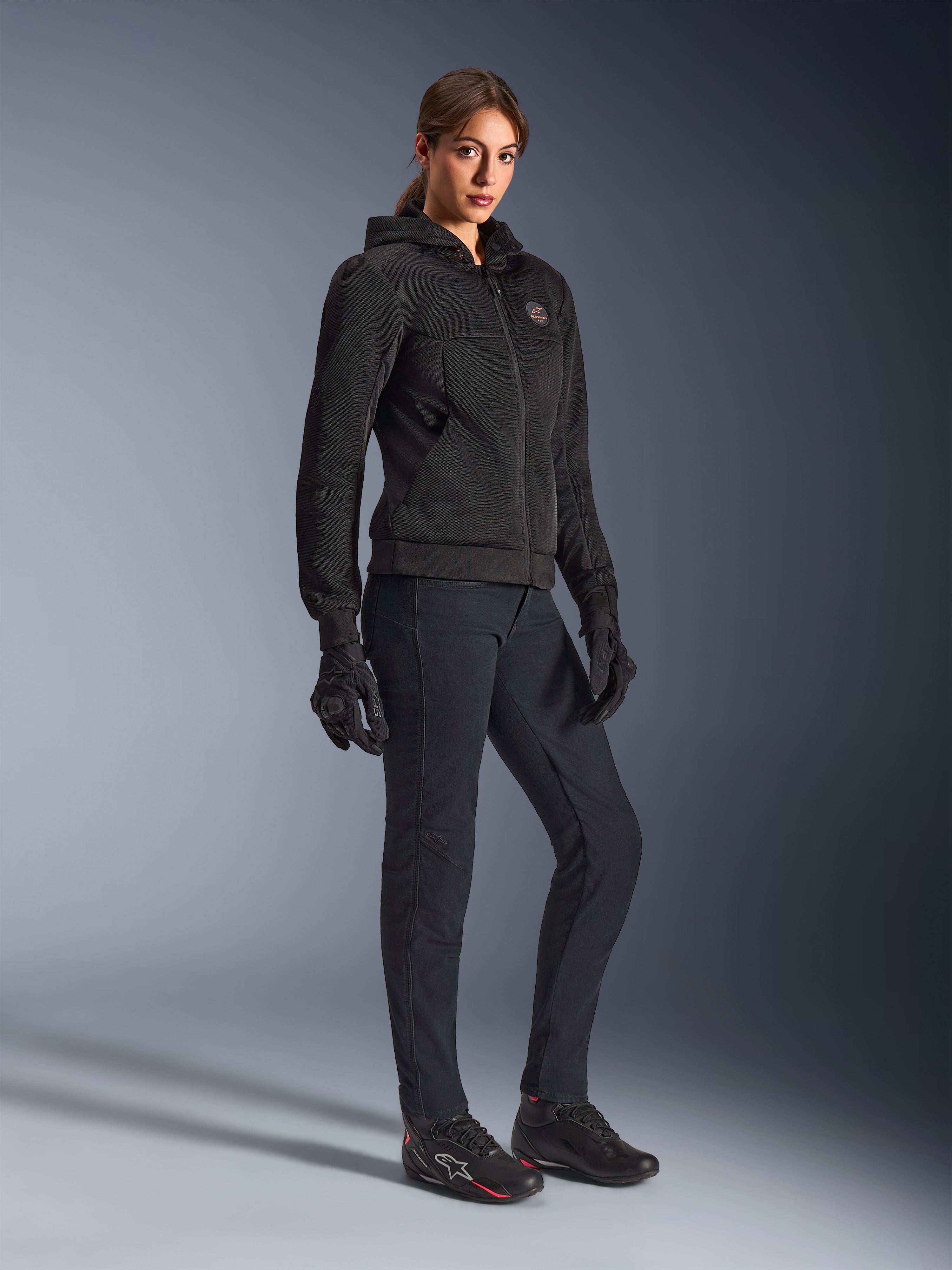 Motocyklistka ubrana w czarną bluzę Alpinestars Stella Chrome Superairflow, techniczna bluza motocyklowa o konstrukcji z siatki 3D, w zestawieniu z czarnymi spodniami motocyklowymi, rękawicami i butami motocyklowymi w sportowym stylu, miejska sportowa estetyka.