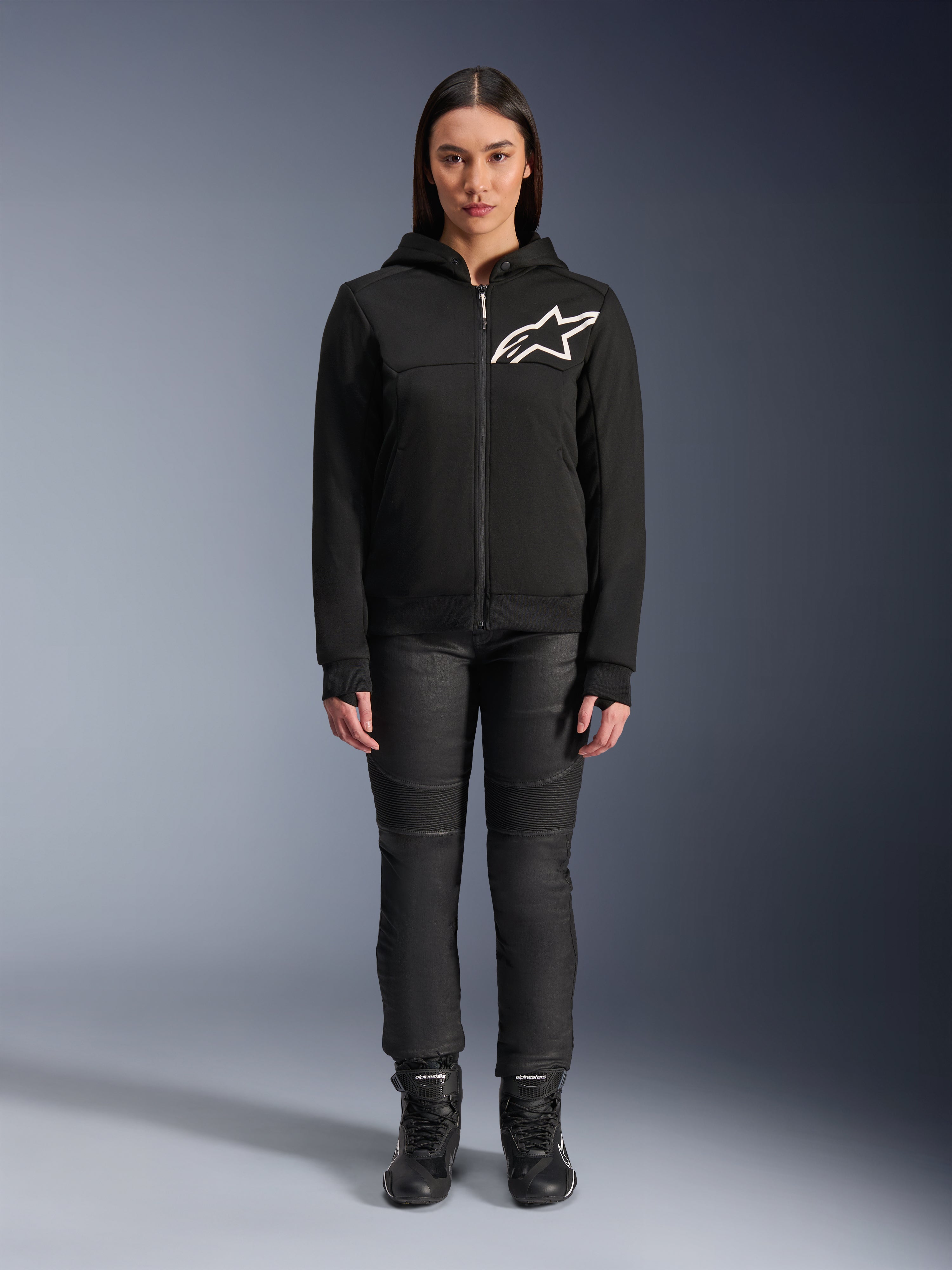 Kobieta w bluzie z kapturem Alpinestars Stella Chrome V2 Sport Hoodie, czarna z białym logo Astars na klatce piersiowej, zestawiona z czarnymi technicznymi spodniami motocyklowymi i butami, miejski styl odzieży motocyklowej.