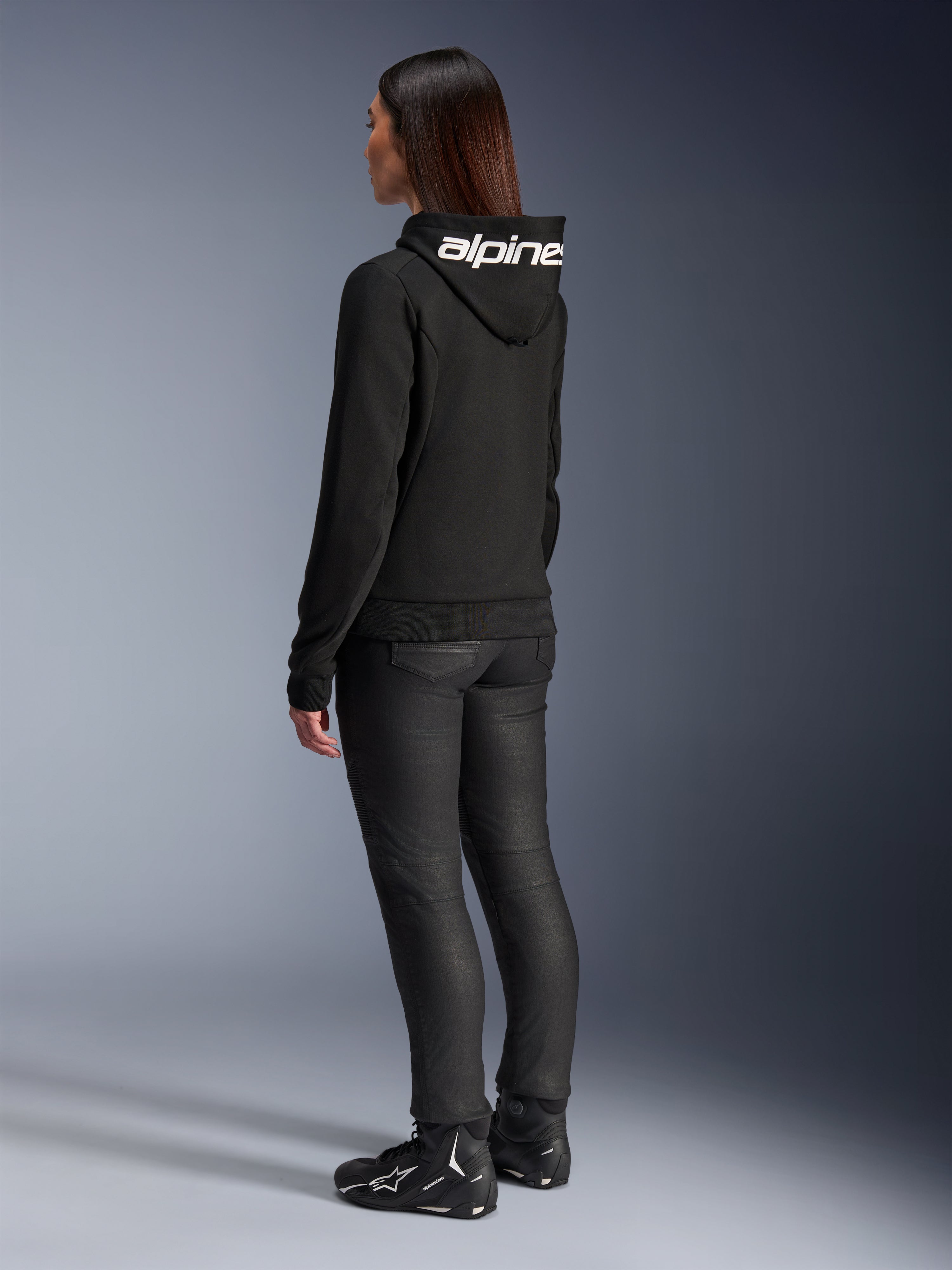 Kobieta ubrana w bluzę Alpinestars Stella Chrome V2 Sport Hoodie, widok z tyłu, czarna motocyklowa bluza z kapturem z białym logo Alpinestars na kapturze, zestawiona z czarnymi technicznymi spodniami do jazdy i czarnymi butami motocyklowymi, casualowy miejski strój do dojazdów