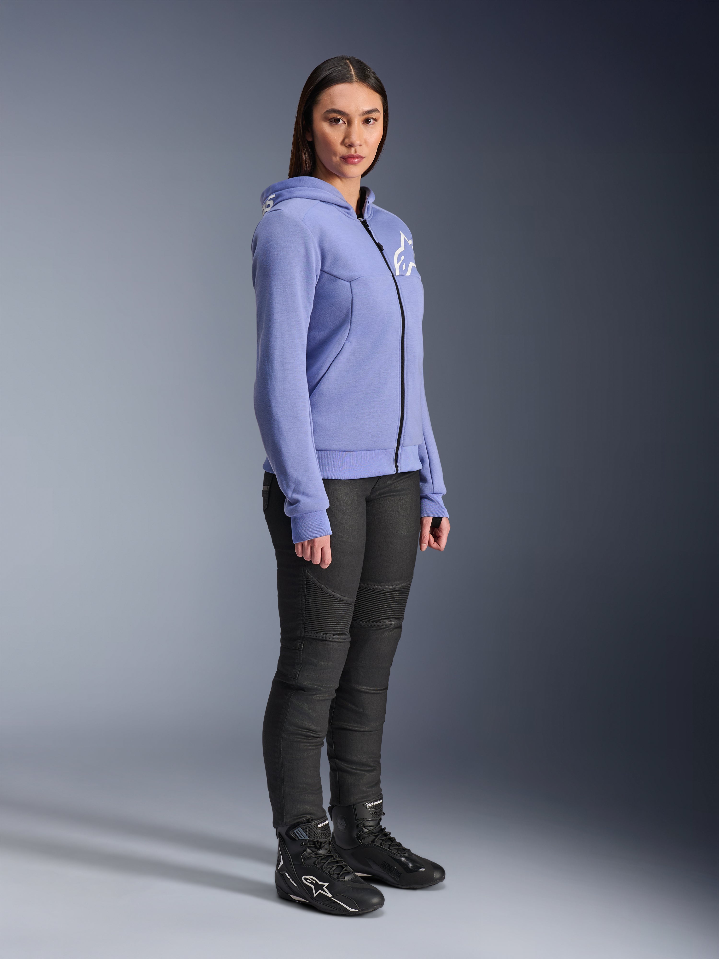 Alpinestars Stella Chrome V2 Sport Hoodie, damska bluza motocyklowa w kolorze niebiesko-fioletowym z białym logo Alpinestars na piersi, prezentowana na modelce z czarnymi spodniami i butami motocyklowymi, miejska odzież motocyklowa dla kobiet