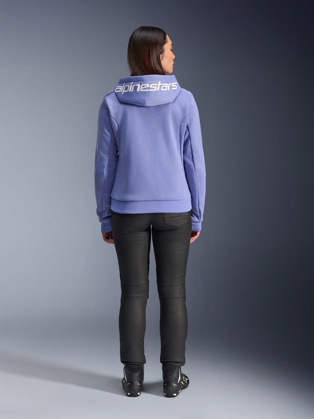 Alpinestars Stella Chrome V2 Sport Hoodie, niebiesko-fioletowa, widok z tyłu kobiety w bluzie z białym logo Alpinestars na kapturze, w zestawieniu z czarnymi spodniami i butami motocyklowymi, codzienna miejska odzież motocyklowa