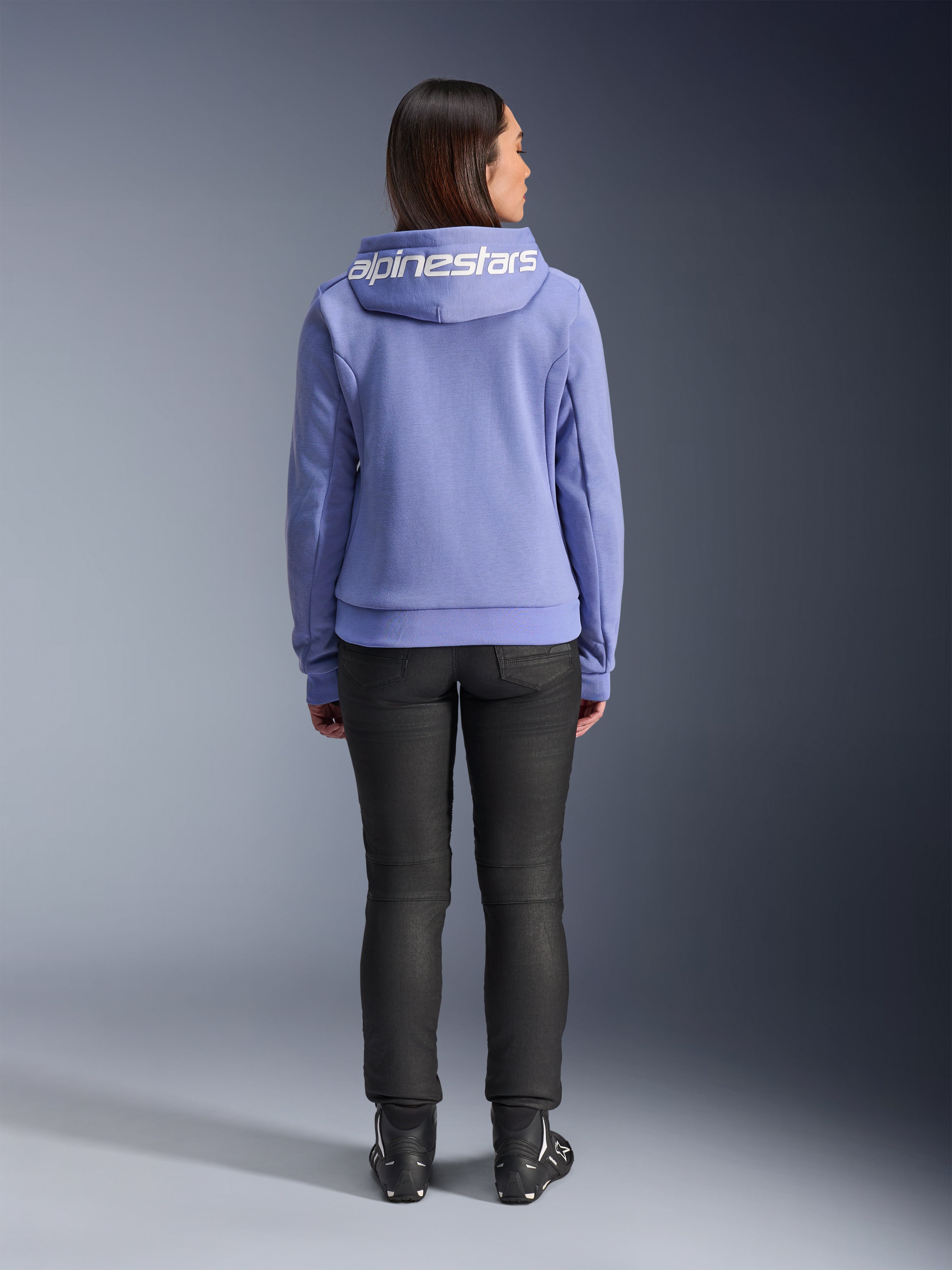 Alpinestars Stella Chrome V2 Sport Hoodie, niebiesko-fioletowa, widok z tyłu kobiety w bluzie z białym logo Alpinestars na kapturze, w zestawieniu z czarnymi spodniami i butami motocyklowymi, codzienna miejska odzież motocyklowa