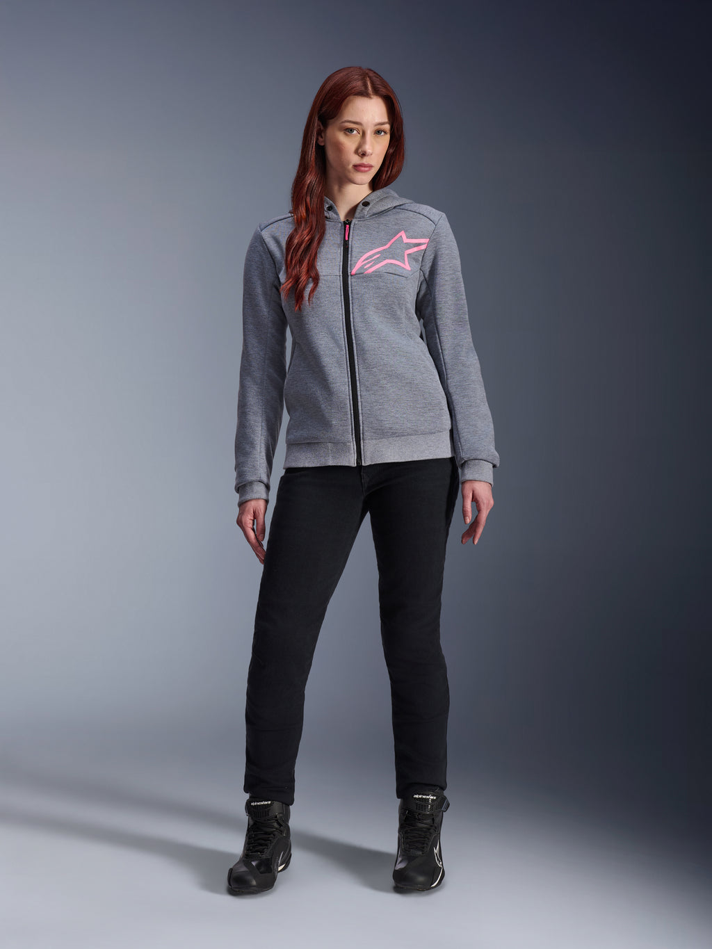 Alpinestars Stella Chrome V2 Sport Hoodie, kolor szary melanż z neonowym różem, noszona przez kobietę w czarnych spodniach i butach, różowe logo gwiazdy Alpinestars na piersi, miejska odzież motocyklowa z pełnym zamkiem, lekki polar techniczny o sportowym kroju.