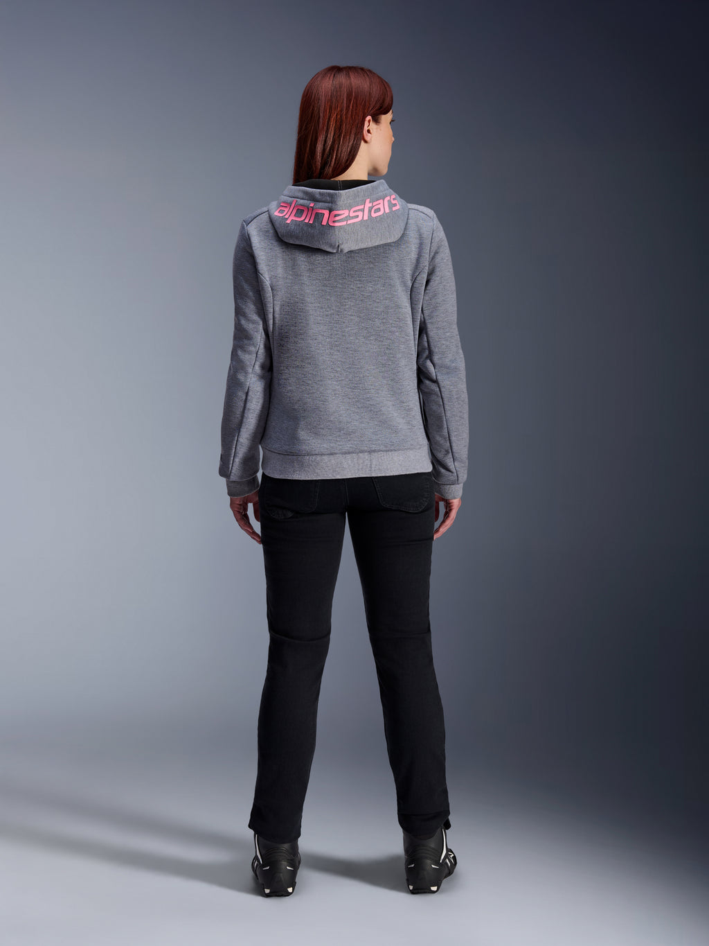 Alpinestars Stella Chrome V2 Sport Hoodie, damska bluza motocyklowa, kolor szary melanż i różowy fluo, widok z tyłu prezentujący różowe logo Alpinestars na kapturze, noszona z czarnymi spodniami i butami motocyklowymi, codzienna odzież motocyklowa dla kobiet
