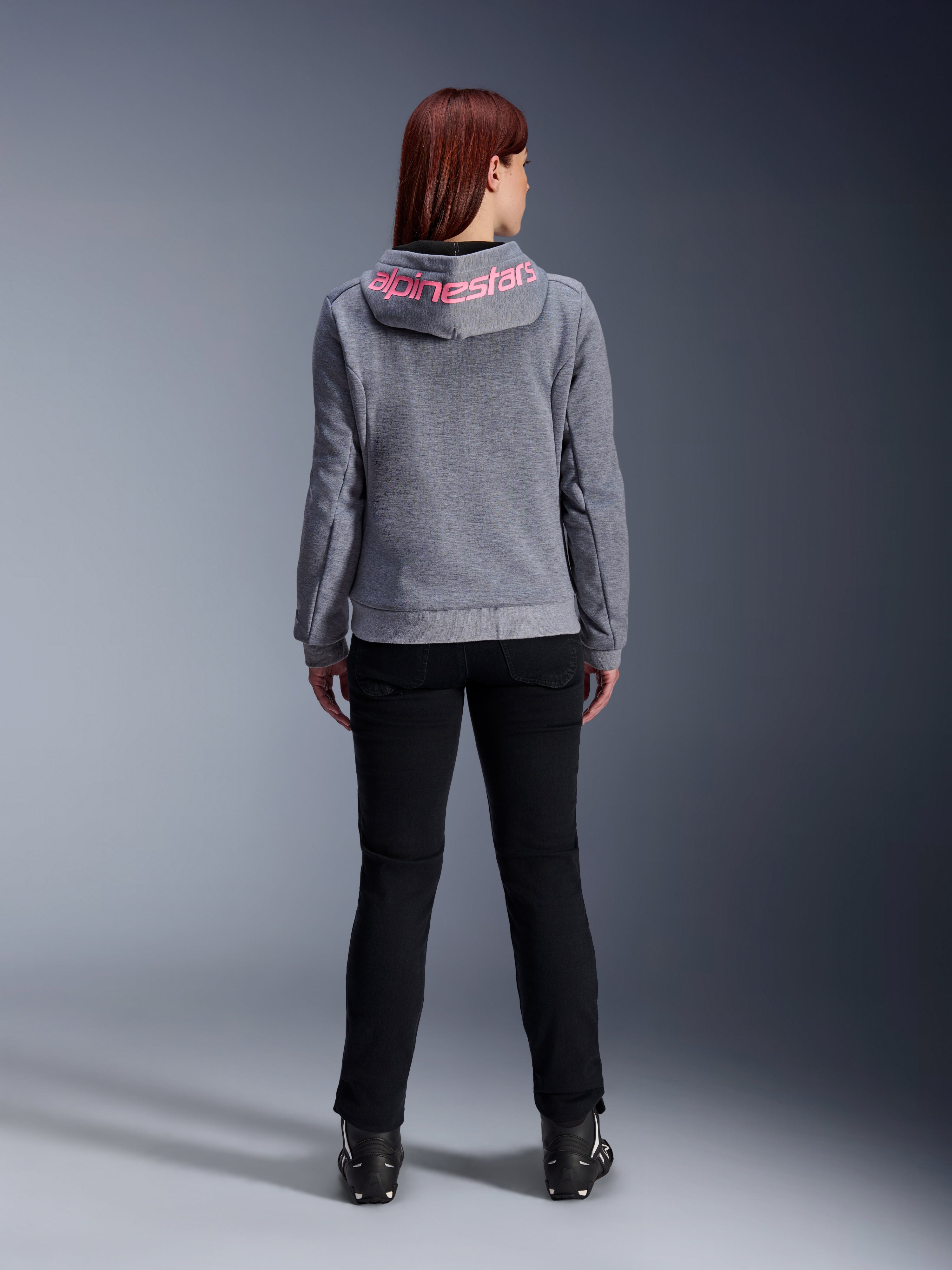 Alpinestars Stella Chrome V2 Sport Hoodie, damska bluza motocyklowa, kolor szary melanż i różowy fluo, widok z tyłu prezentujący różowe logo Alpinestars na kapturze, noszona z czarnymi spodniami i butami motocyklowymi, codzienna odzież motocyklowa dla kobiet