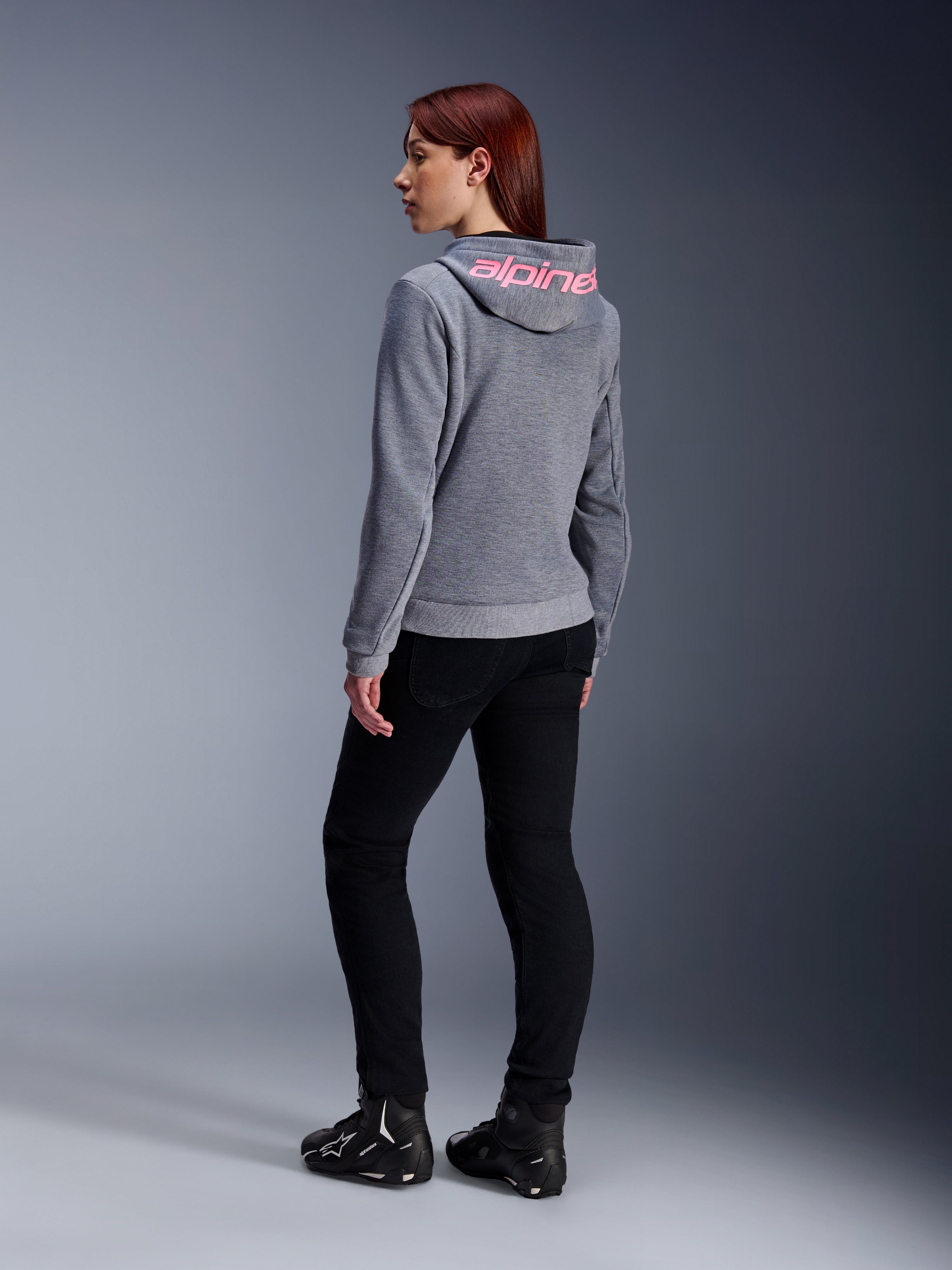 Alpinestars Stella Chrome V2 Sport Hoodie, damska bluza motocyklowa, szary melanż z różowym neonowym brandingiem na kapturze, widok z tyłu, zestawiona z czarnymi spodniami i butami motocyklowymi, swobodny miejski styl z ochroną motocyklową