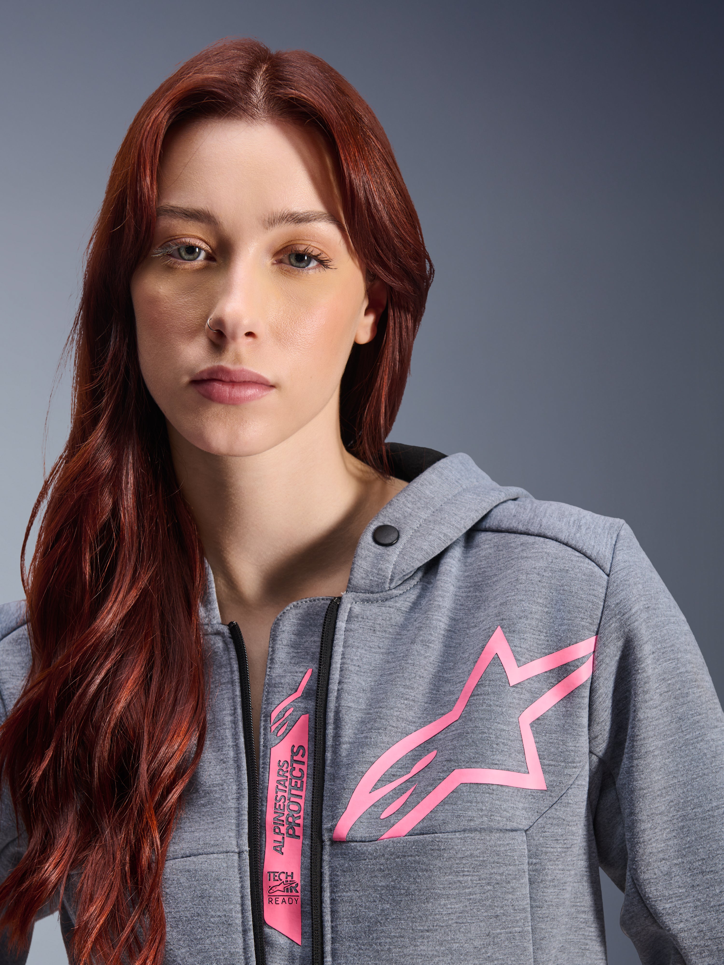 Kobieta w bluzie Alpinestars Stella Chrome V2 Sport Hoodie, Gray Heather Pink Fluo, zbliżenie na różowe logo gwiazdy Alpinestars oraz oznaczenie Tech-Air Ready, miejska odzież motocyklowa