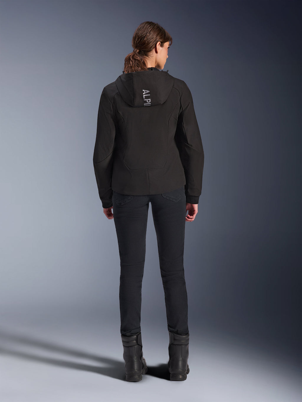 Alpinestars Woman Stella Radium Tech Hoodie, czarna, widok z tyłu na modelkę ubraną w lekką, 3-warstwową wiatroszczelną bluzę techniczną z logo Alpinestars na kapturze, w zestawie z czarnymi spodniami i butami, miejska odzież motocyklowa