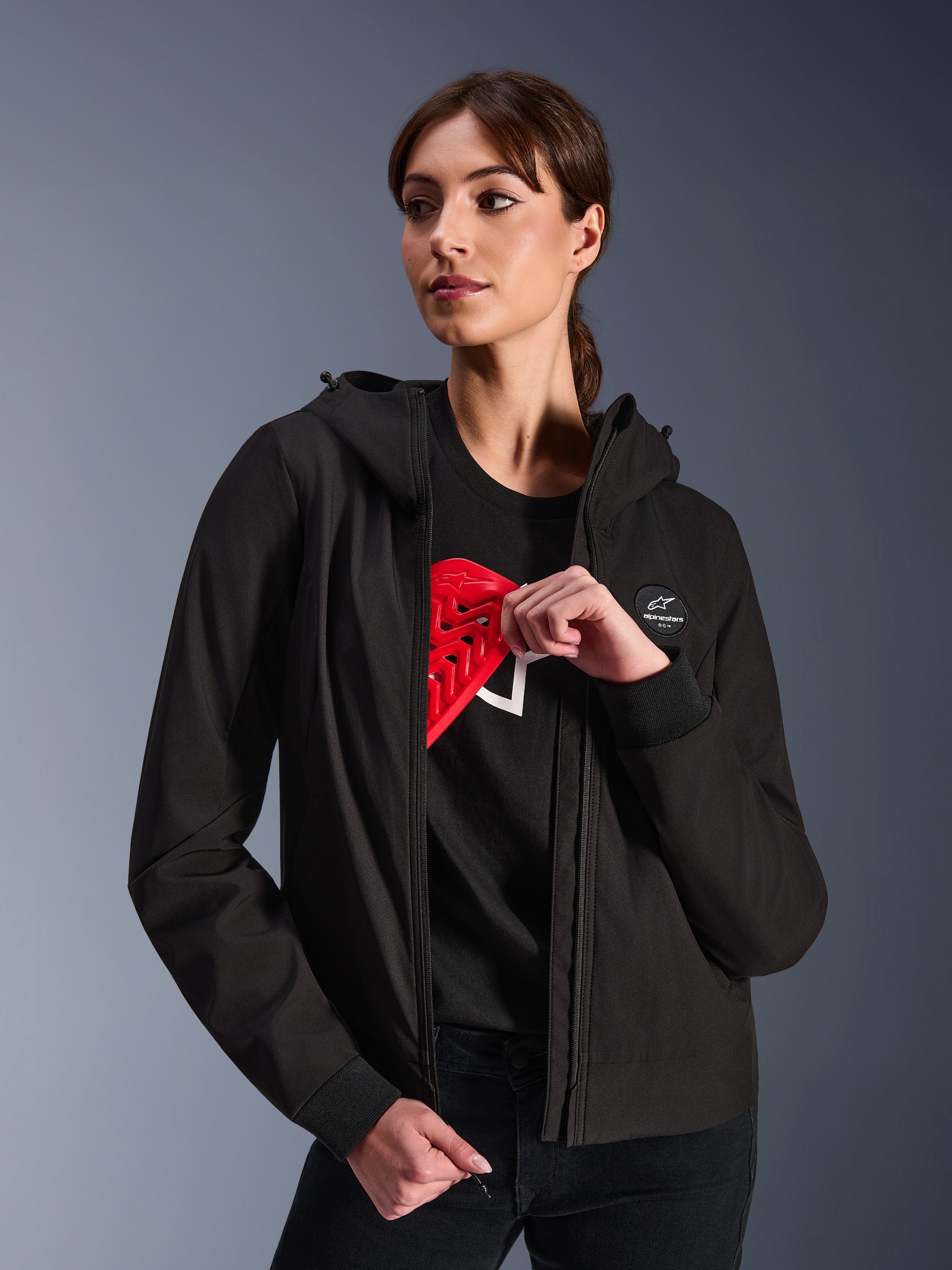 Alpinestars Woman Stella Radium Tech Hoodie, czarna miejska bluza motocyklowa z kapturem, modelka prezentująca czerwony ochraniacz Nucleon Flex Plus wewnątrz, lekka 3-warstwowa wiatroszczelna powłoka tekstylna ze ściągaczami i naszywką z logo na piersi, ochronna odzież motocyklowa