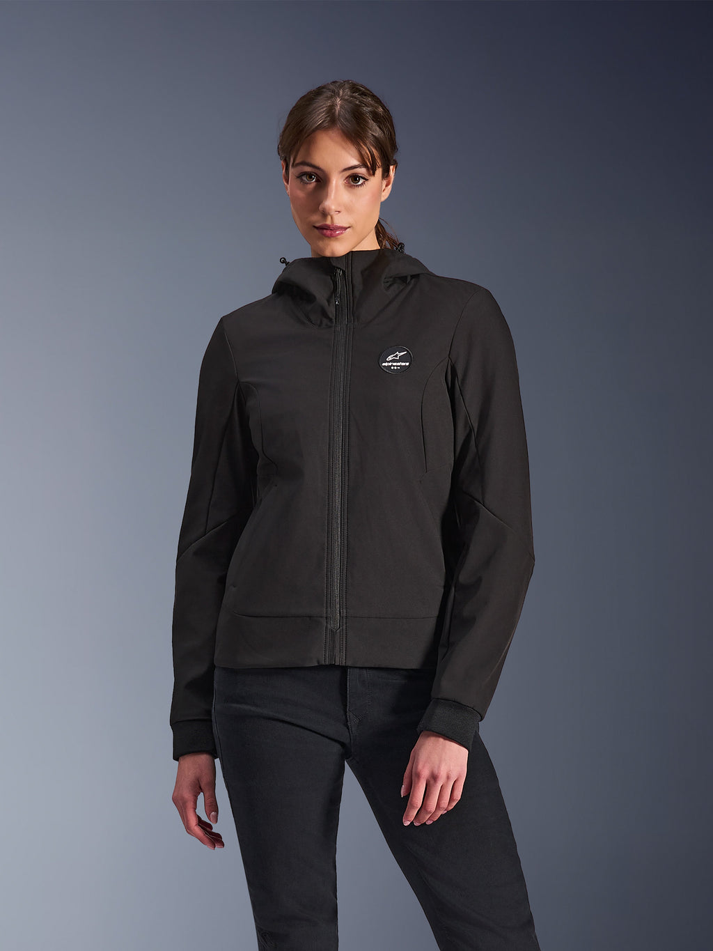 Alpinestars Stella Radium Tech Hoodie, damska motocyklowa bluza z kapturem, czarna, fason zapinany na zamek z okrągłym logo na piersi i kapturem, techniczna odzież do jazdy miejskiej, prezentowana przez modelkę