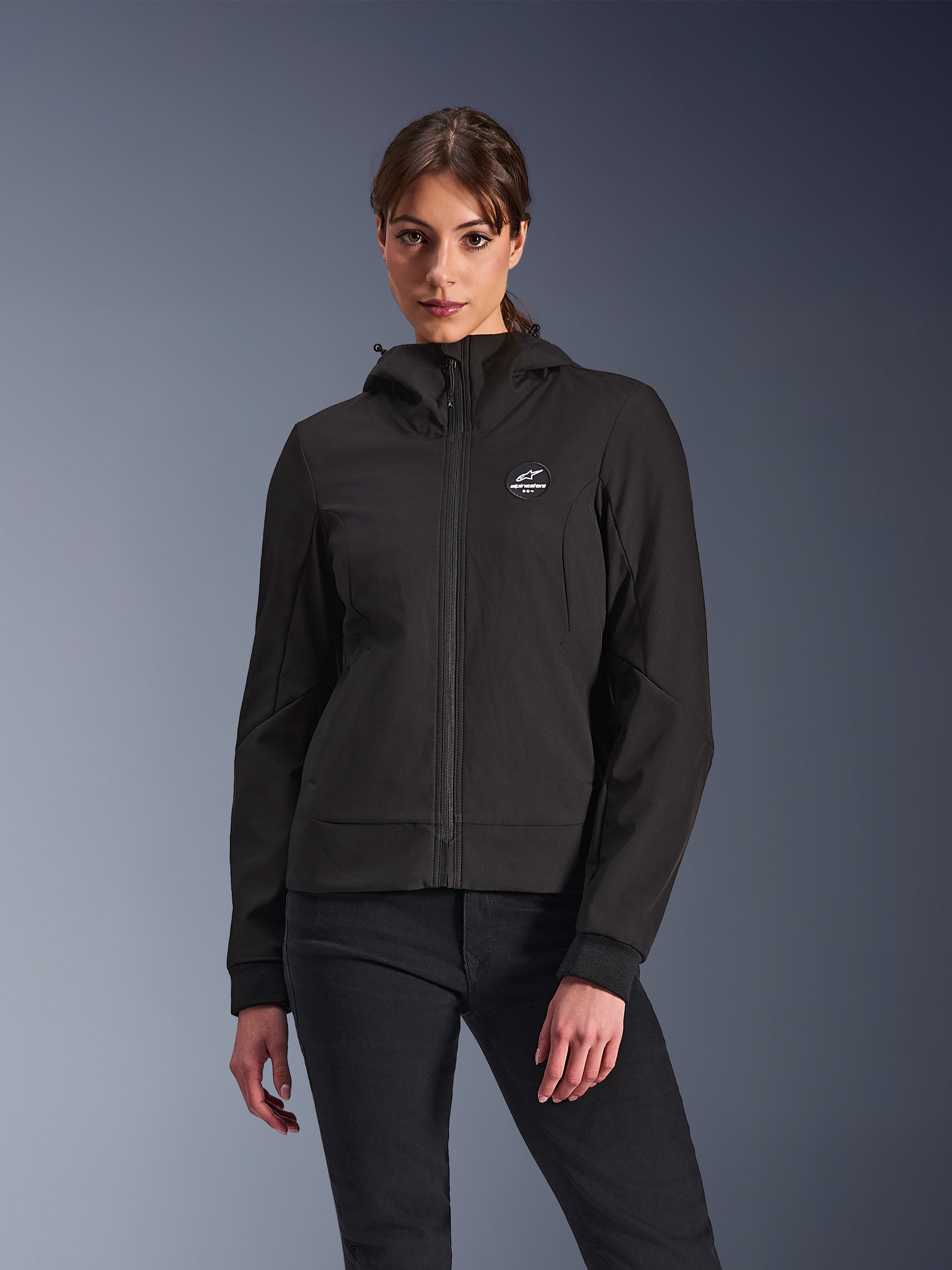 Alpinestars Stella Radium Tech Hoodie, damska motocyklowa bluza z kapturem, czarna, fason zapinany na zamek z okrągłym logo na piersi i kapturem, techniczna odzież do jazdy miejskiej, prezentowana przez modelkę