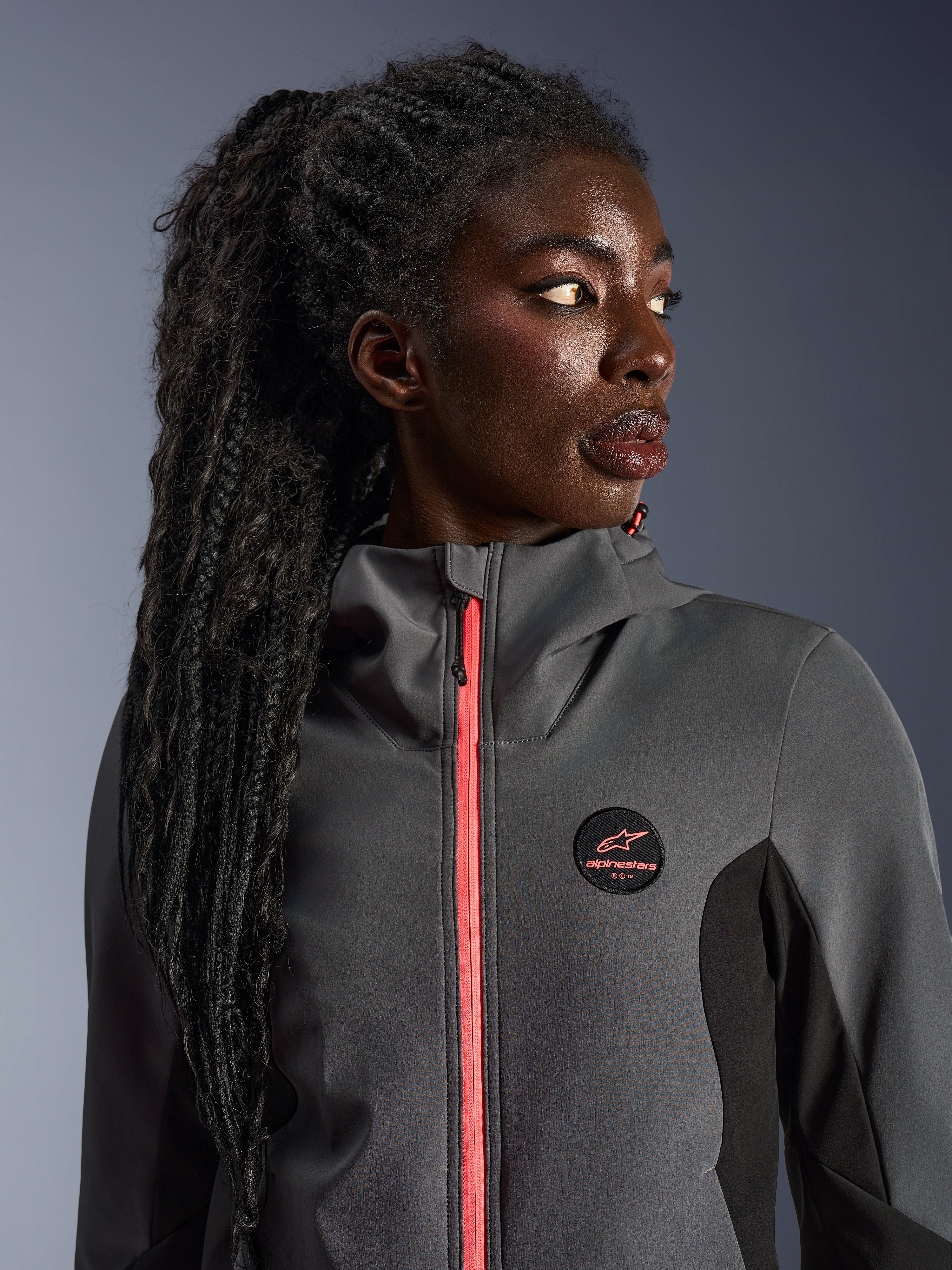 Modelka w bluzie Alpinestars Stella Radium Tech Hoodie w kolorze ciemnoszarym i czarnym, z jaskrawym koralowym zamkiem, okrągłym logo na piersi i wiatroszczelną powłoką z wysokim kołnierzem, miejska odzież motocyklowa dla kobiet