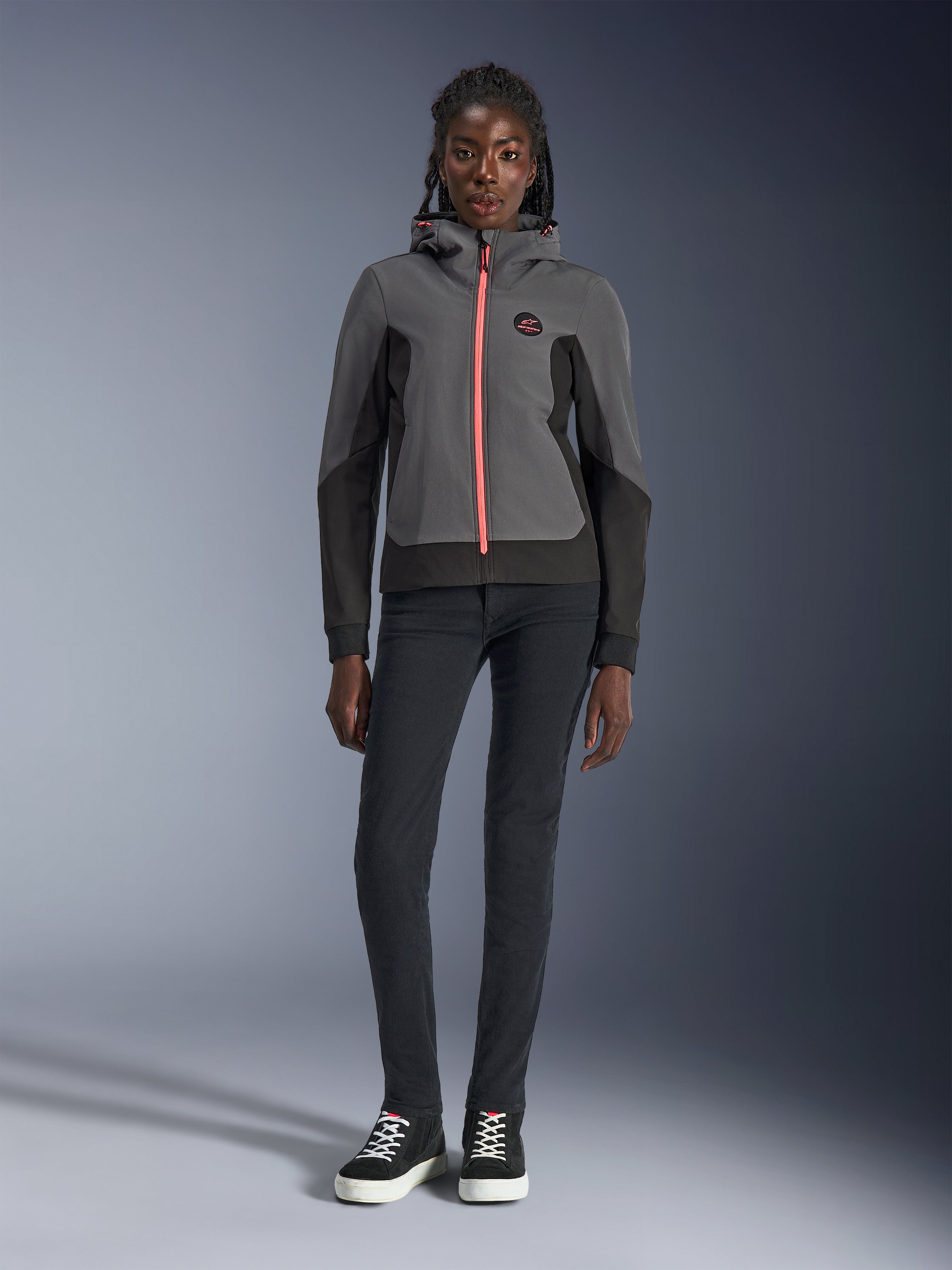 Modelka w bluzie Alpinestars Stella Radium Tech Hoodie, kolor ciemnoszary i czarny z jaskrawopomarańczowym akcentem zamka, miejska bluza motocyklowa, w zestawieniu z czarnymi spodniami typu slim i sneakersami, casualowa odzież sportowa dla kobiet