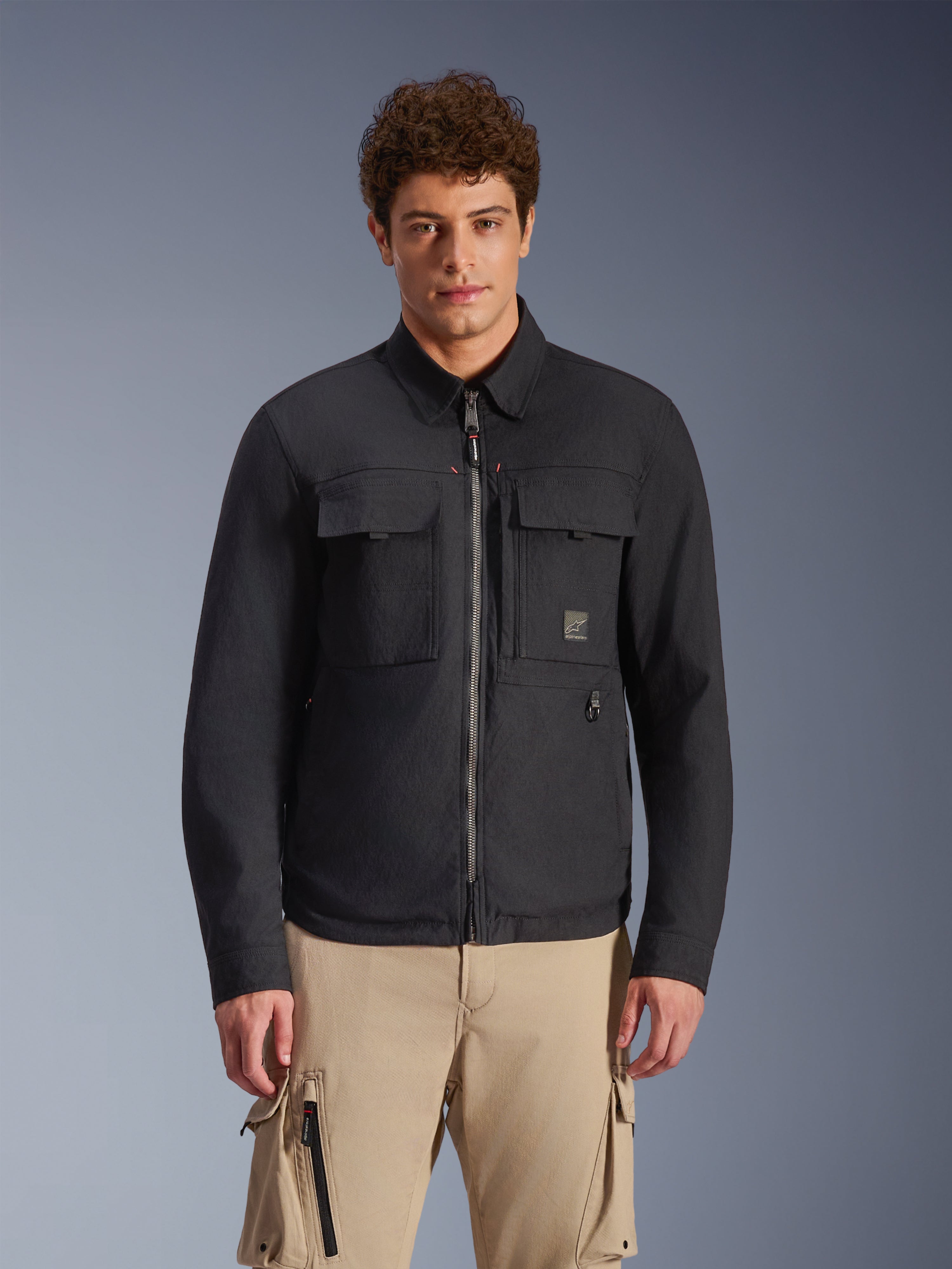 Alpinestars Bryon Water Resistant Overshirt, czarna miejska kurtka motocyklowa w stylu roboczym, dwie duże kieszenie cargo na piersi, zamek błyskawiczny na całej długości, wykonana z woskowanego płótna, model w beżowych spodniach cargo.