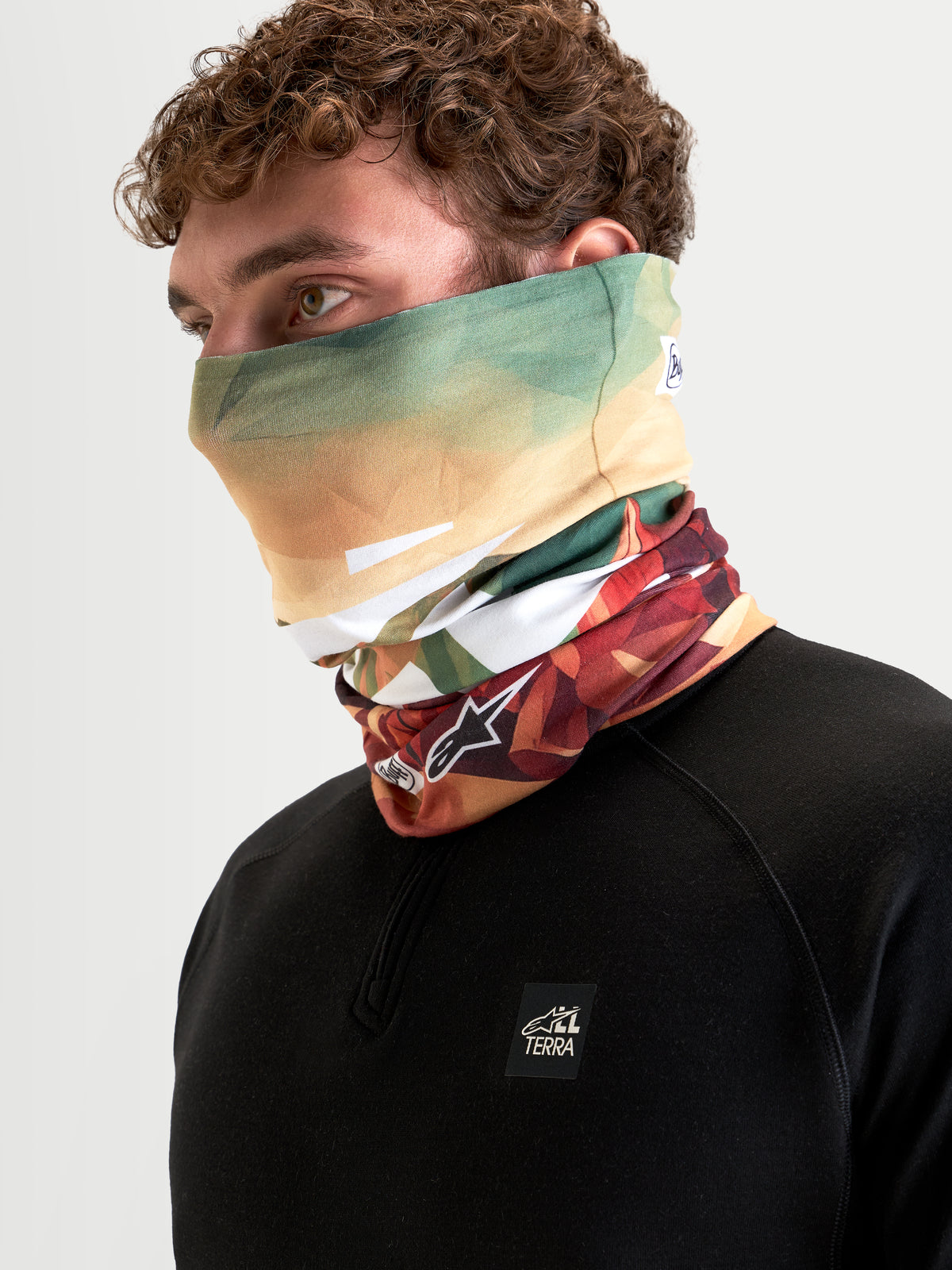 Alpinestars Buff Peaks PrimaLoft Neck Tube, motorcycle neck gaiter in Earth Brown and green, worn by a model, featuring moisture-wicking and low-bulk insulated textile construction for cool-weather riding