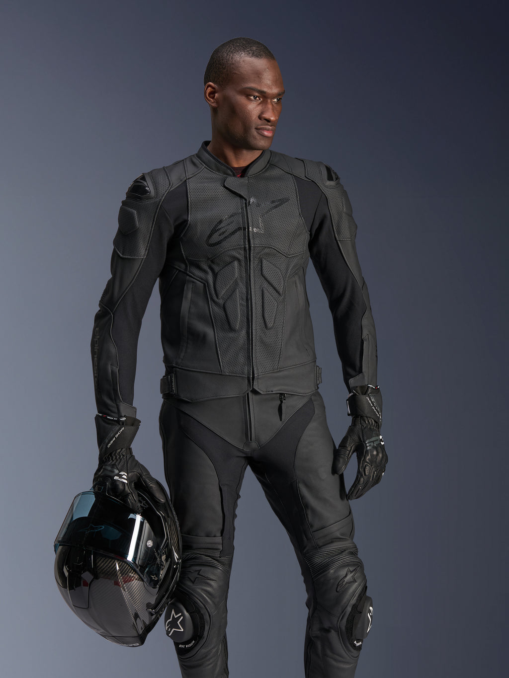 Alpinestars Dusk Airflow Leather Jacket, czarna skórzana kurtka motocyklowa, noszona przez motocyklistę z pasującymi skórzanymi spodniami i rękawicami, trzymającego czarny kask, perforowane panele skórzane, zintegrowane ochraniacze do sportowej jazdy