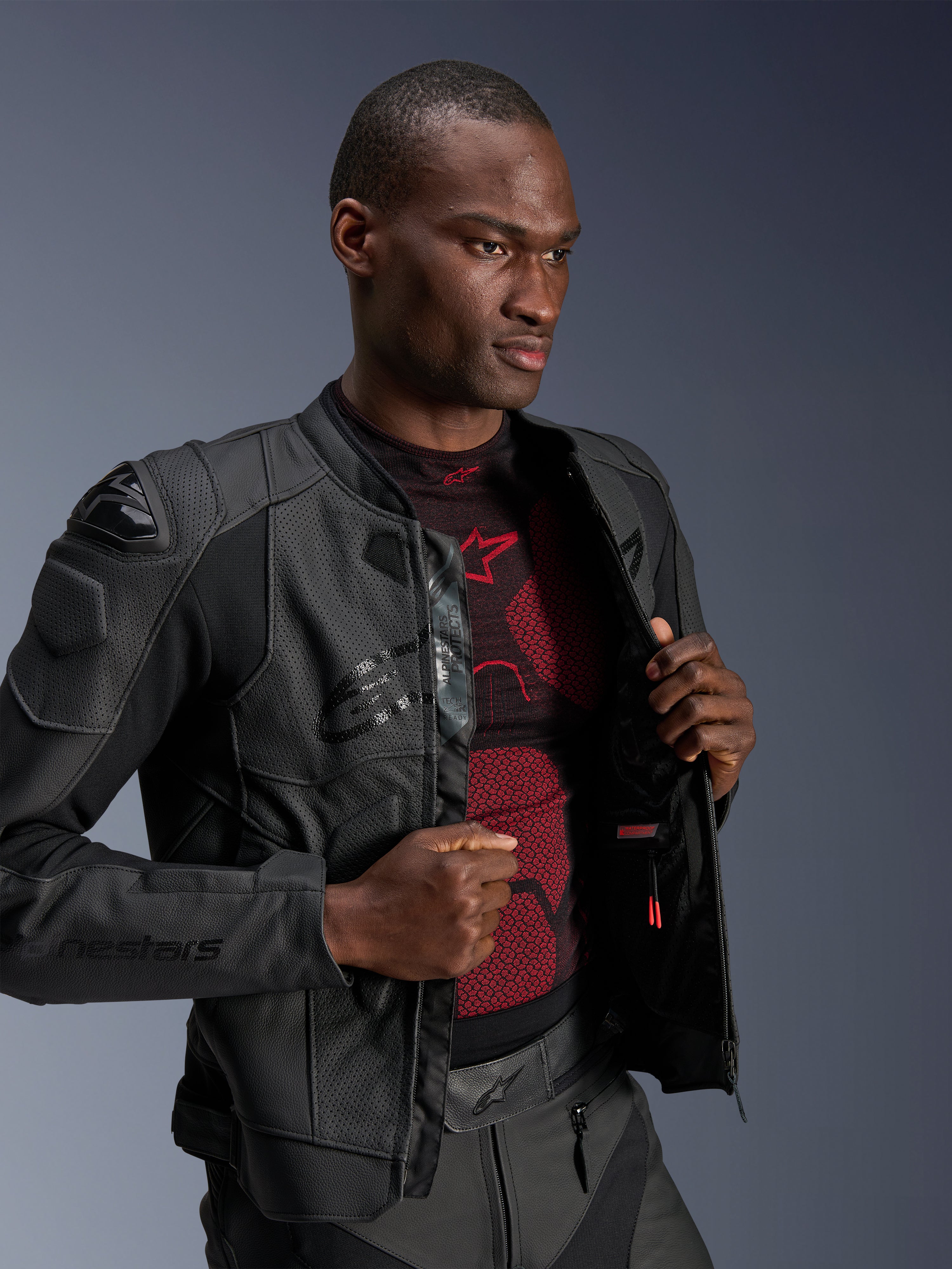 Skórzana kurtka Alpinestars Dusk Airflow, kolor Black Black, noszona przez modela na czerwono-czarnej warstwie bazowej, z perforowanymi panelami skórzanymi, zewnętrznymi sliderami ramion DFS i o sportowym kroju.