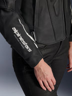 Alpinestars Stella Dusk Leather Jacket, zbliżenie na damską czarną skórzaną kurtkę motocyklową, białe logo „alpinestars RACING AHEAD” na rękawie, tekstura najwyższej jakości skóry bydlęcej, noszona z czarnymi spodniami